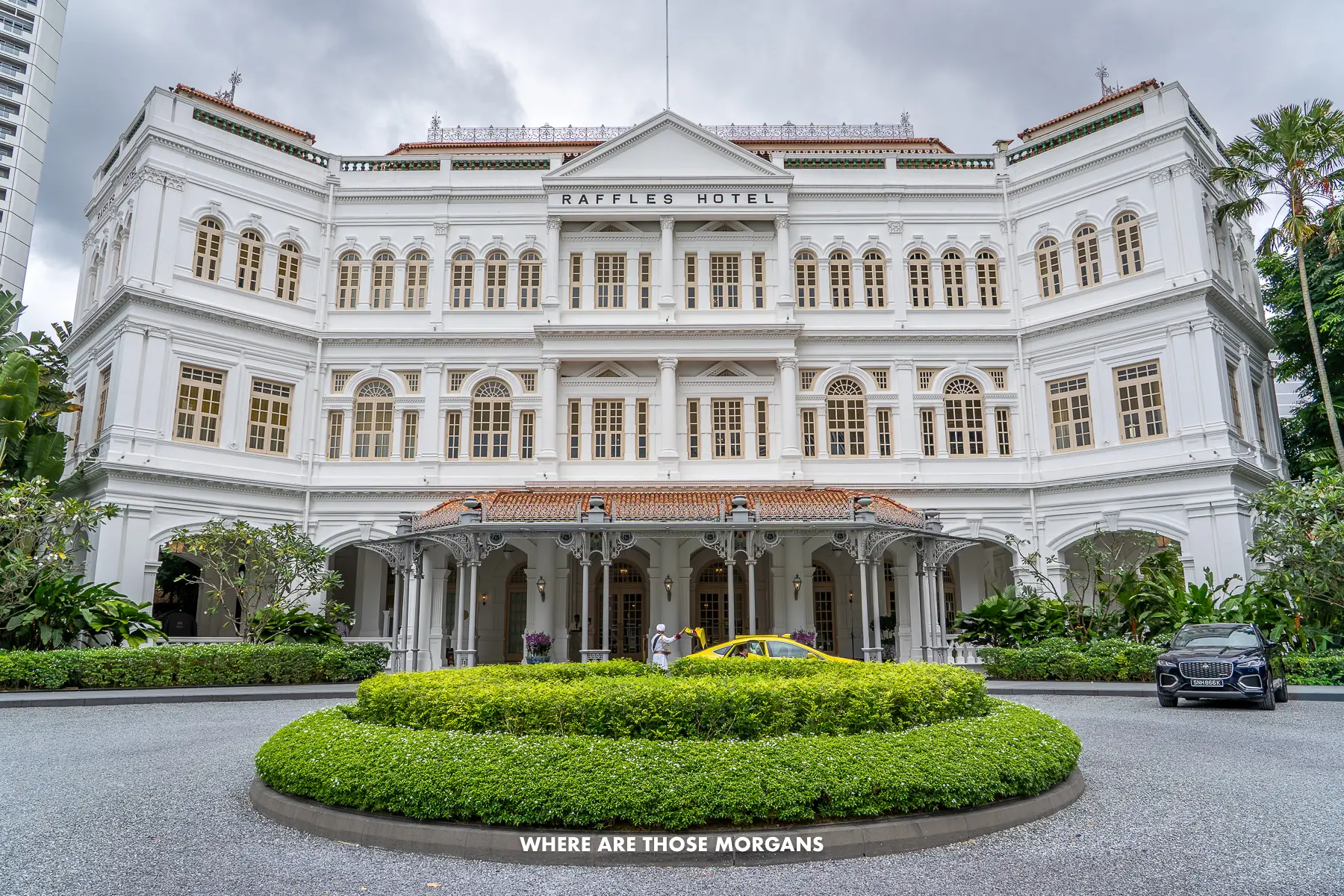 Raffles hotel in Singapore from the front outside with a car parked outside and a taxi being opened by a bellboy