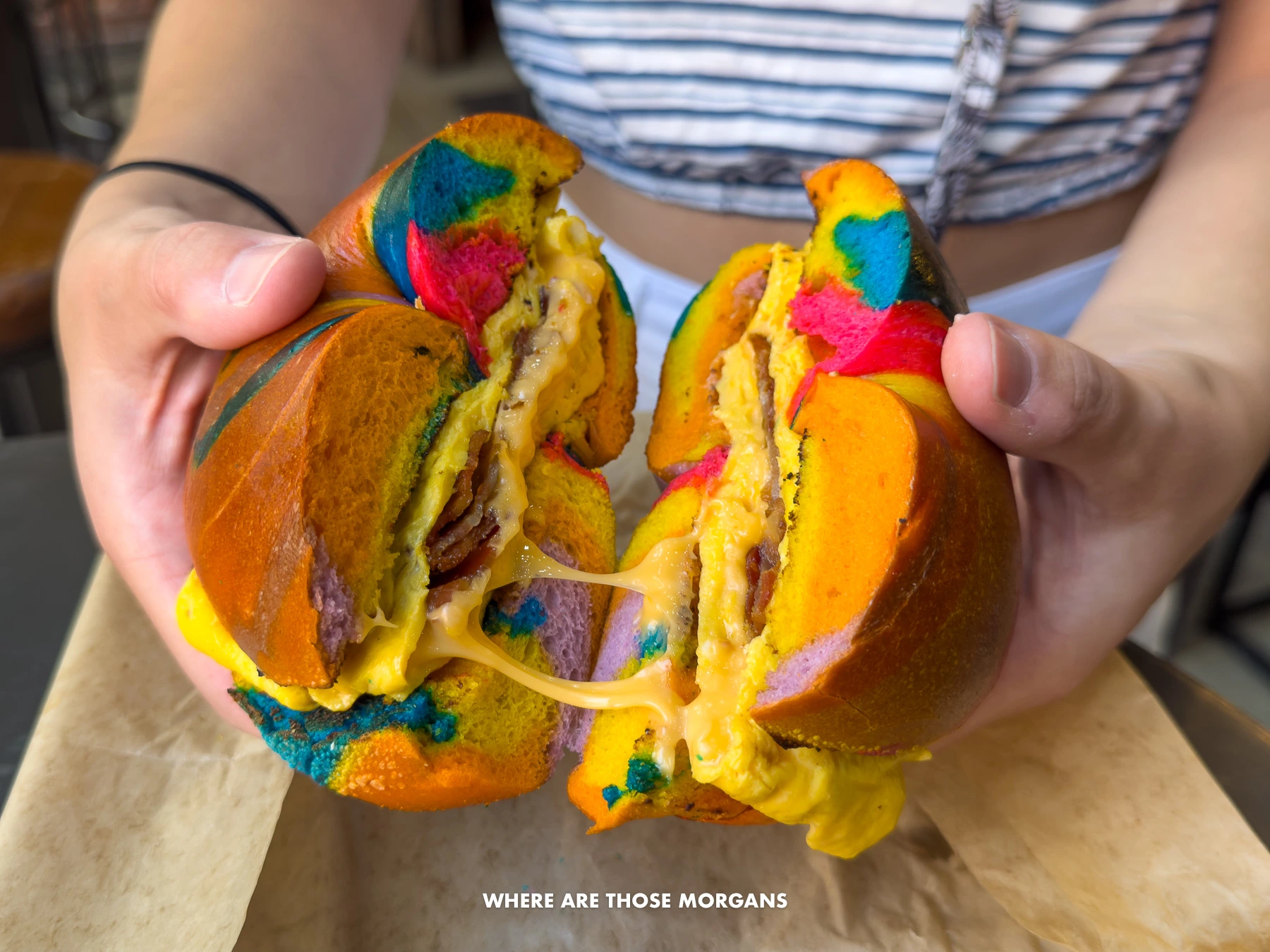 Person holding a rainbow colored bagel that's been cut in half with cheese stretching between the two sides