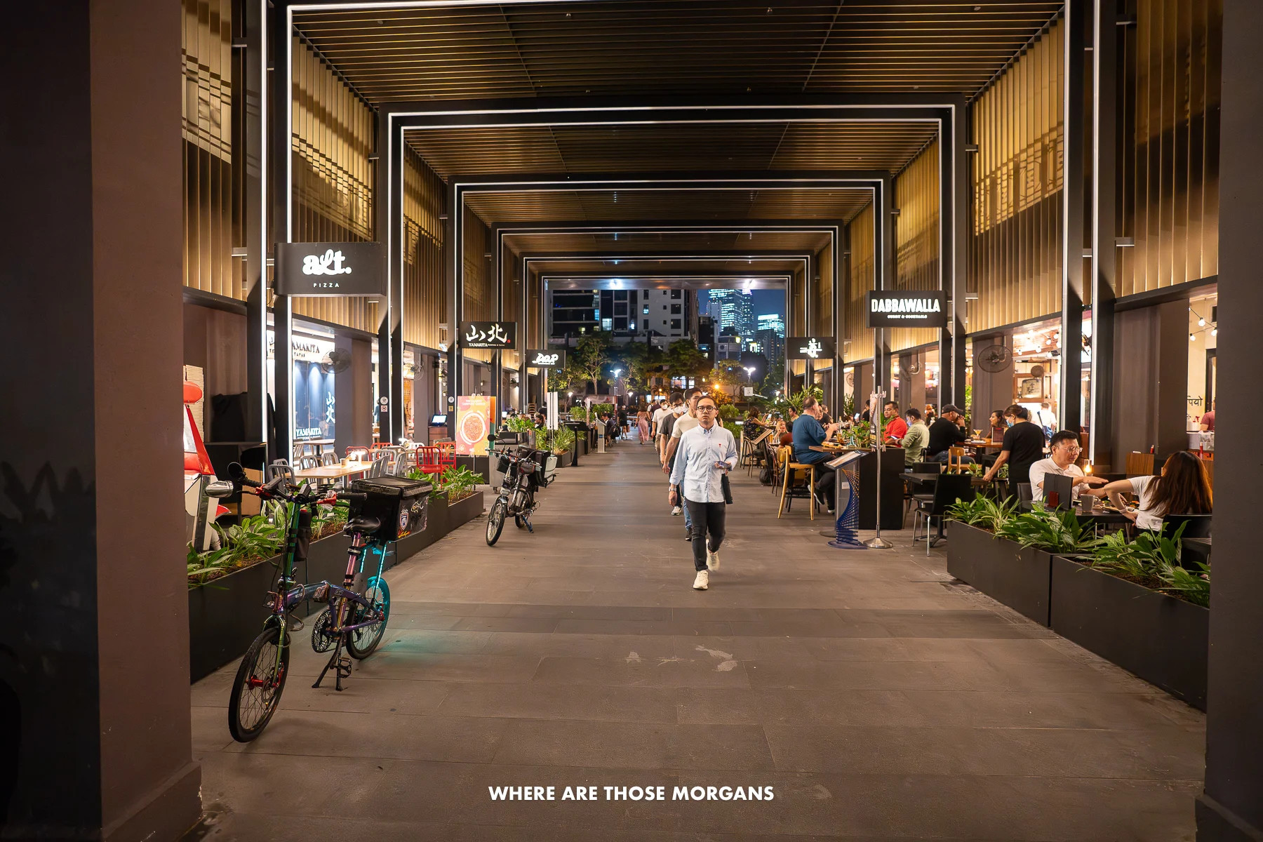 People sat at tables and walking through the underneath of a building with restaurants at night