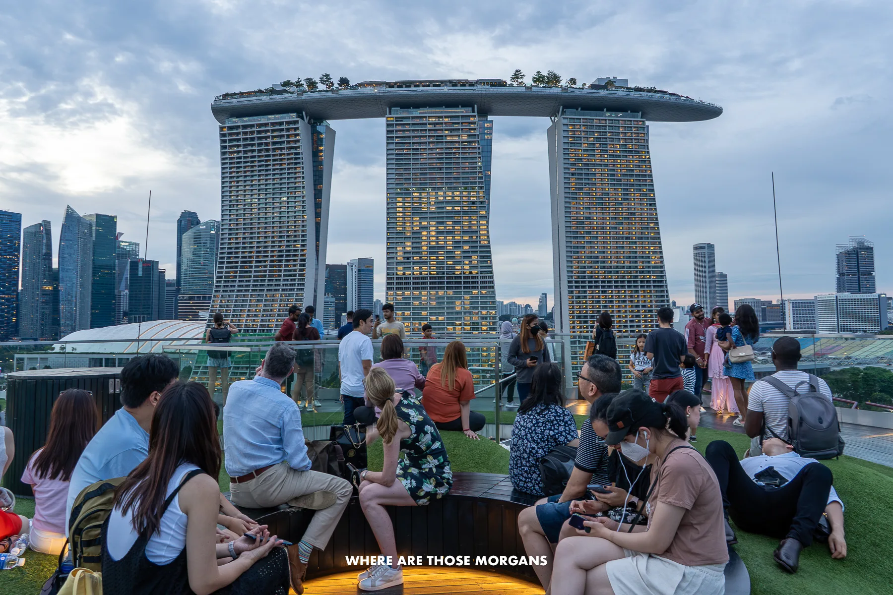 Small crowd of people sat at the top of an observation deck in Gardens by the Bay overlooking the three towers and top of Marina Bay Sands in Singapore at dusk