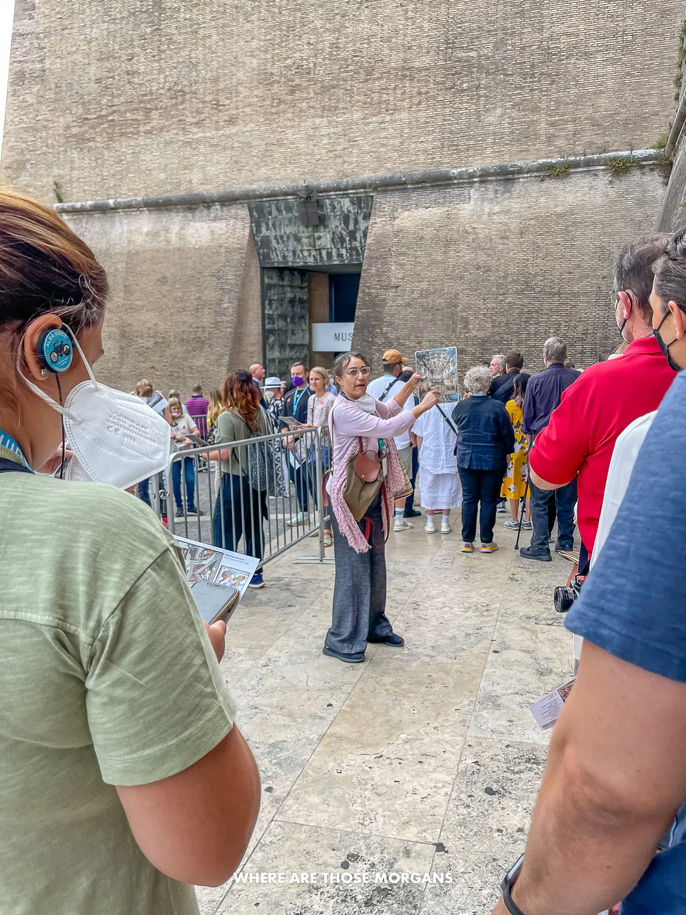 A tour guide explaining things from a laminated sheet to tourists with head sets on at the entrance to a building