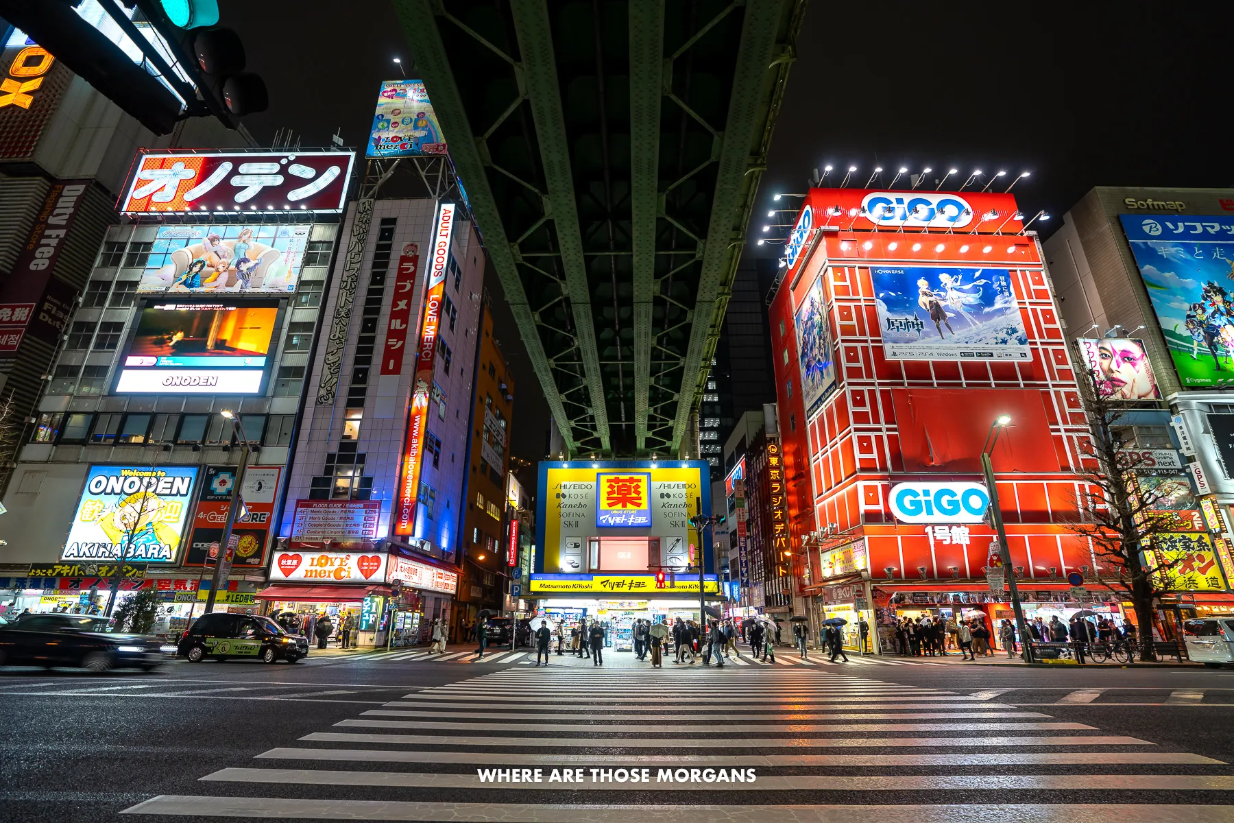Pedestrian crossing under a bridge on a wide open road with arcades in skyscrapers lit up with bright colors at night in Akihabara
