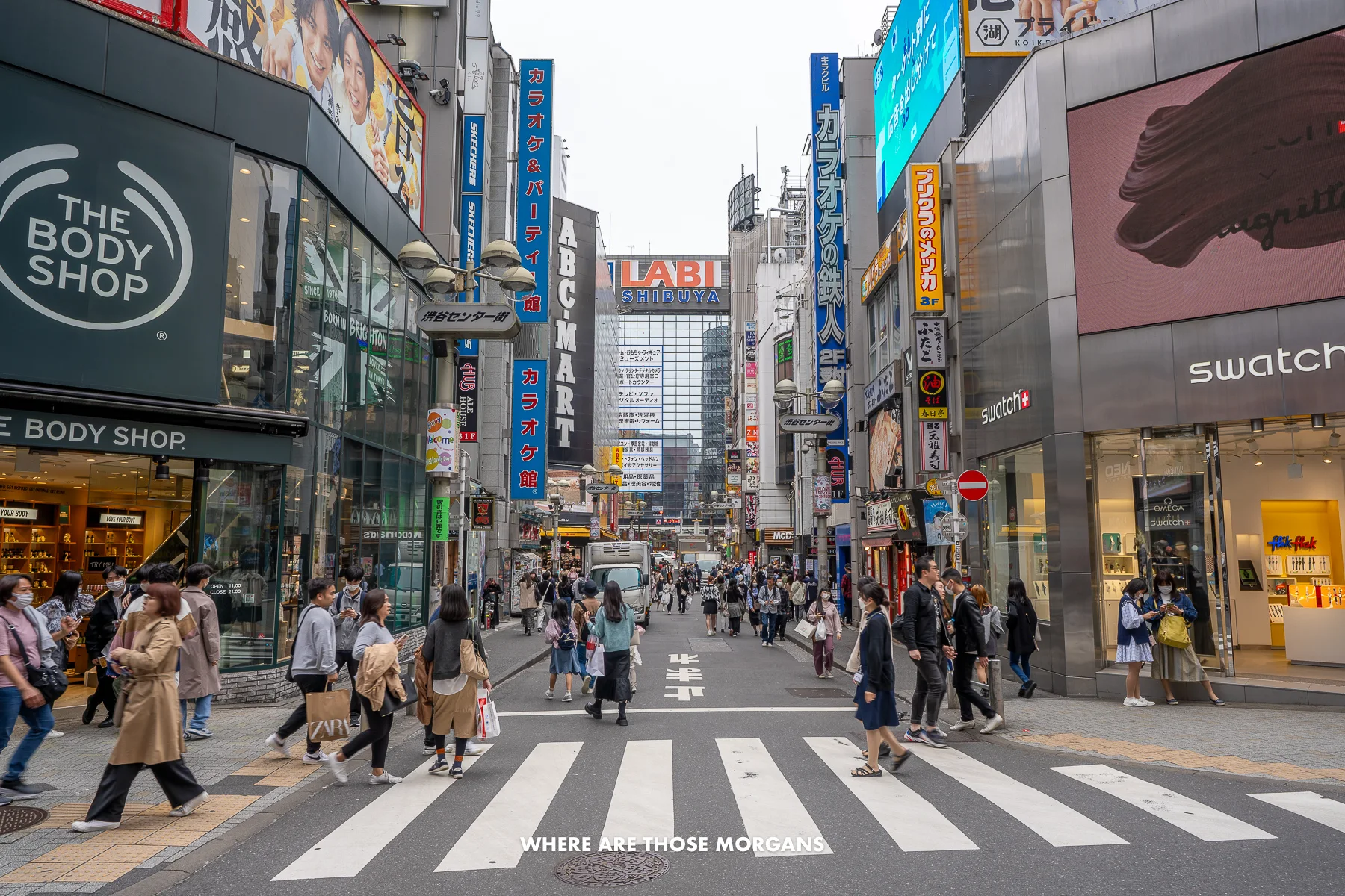 Lots of tourists and locals walking across a road with pedestrian crossing between a row of buildings