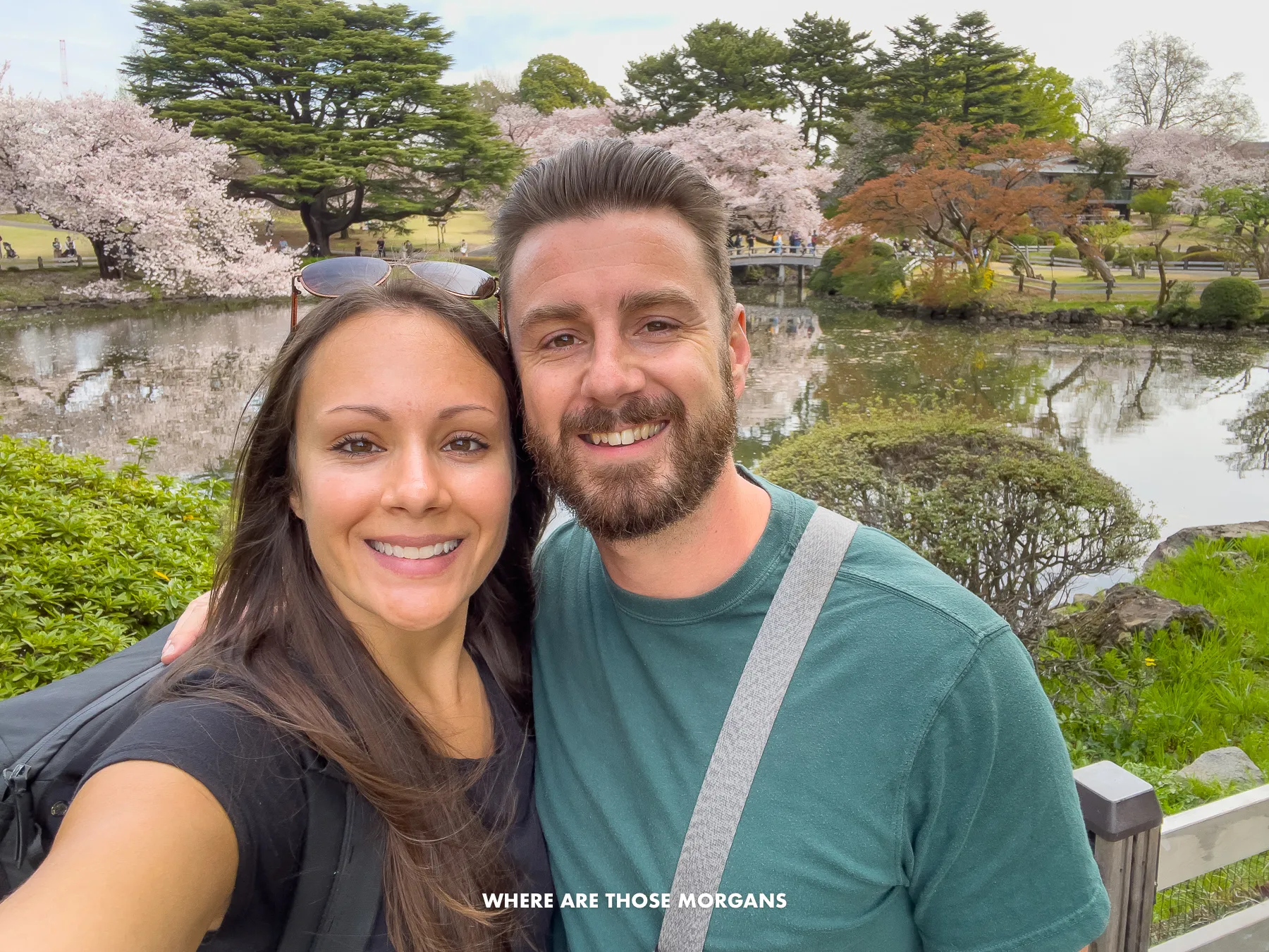 Mark and Kristen from Where Are Those Morgans taking a selfie in Shinjuku Gyeon National Garden in Tokyo during cherry blossom season with a small pond and trees behind
