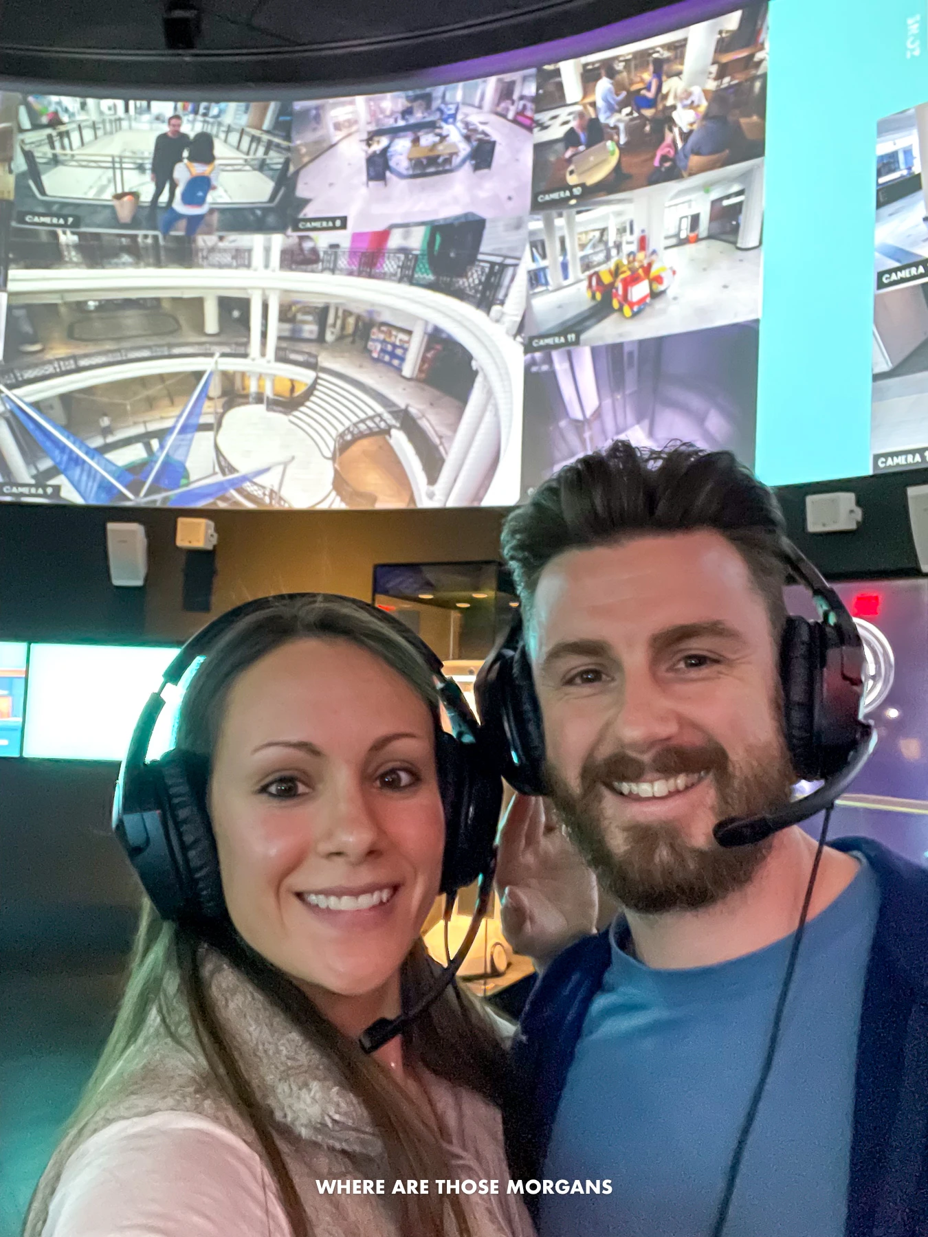 Mark and Kristen from Where Are Those Morgans taking a selfie inside a spy museum in New York City with headsets and microphones on, and a massive tv screen behind