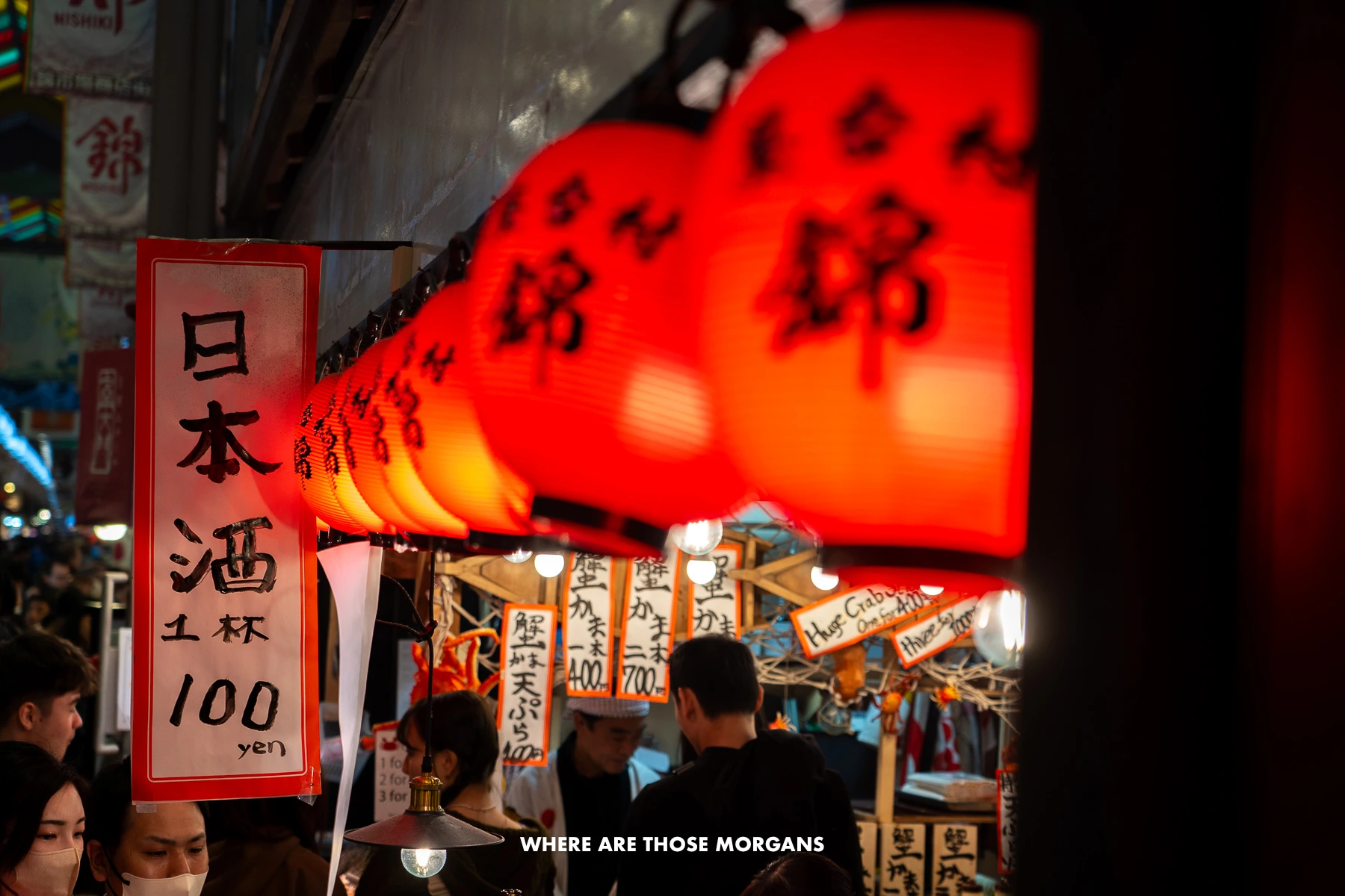 Red lanterns above a food stall inside a market in Kyoto with tourists browsing