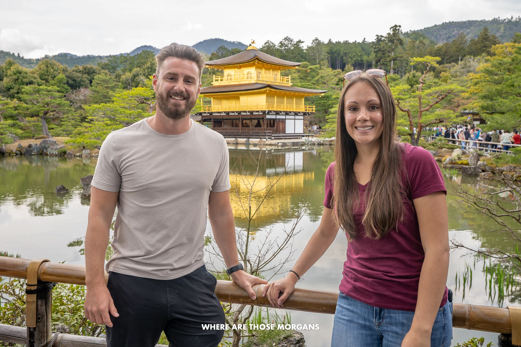 Mark and Kristen from Where Are Those Morgans standing together in front of a low wooden fence with a pond and a golden pavilion reflecting behind on a cloudy day at Kinkaku-ji Temple in Kyoto