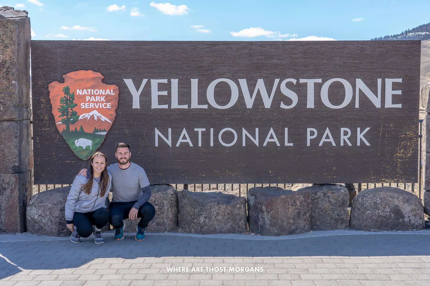 Mark and Kristen from Where Are Those Morgans crouched down together in front of a wooden Yellowstone National Park sign on a road trip from Mt Rushmore