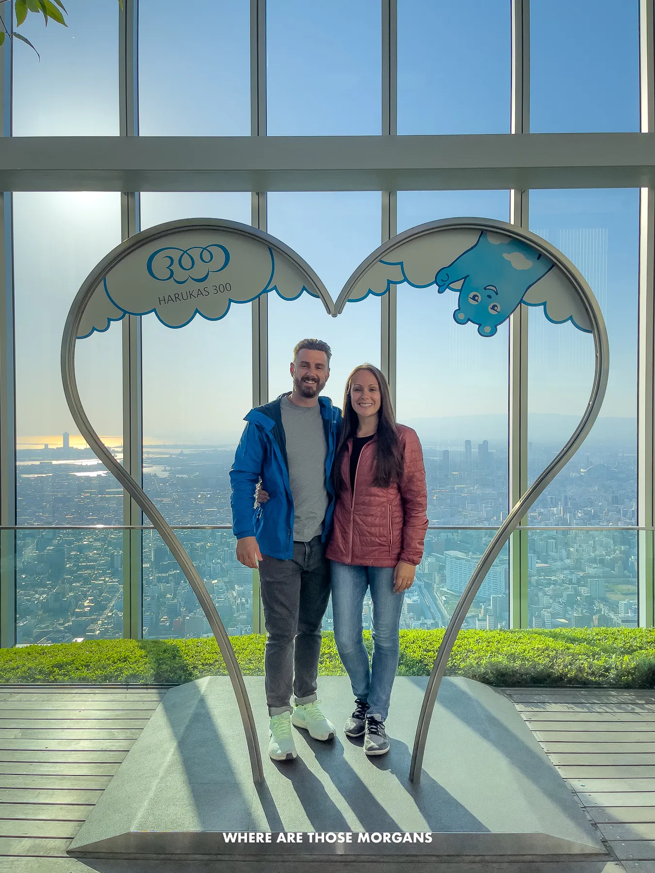 Mark and Kristen from Where Are Those Morgans standing together underneath a love heart shaped exhibit with massive glass windows behind and views over a city in Japan
