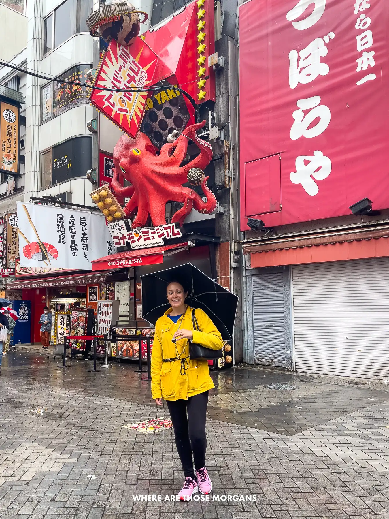 Kristen from Where Are Those Morgans wearing a yellow coat and holding a black umbrella on a rainy day walking through Dotonbori in Osaka with an octopus restaurant facade behind