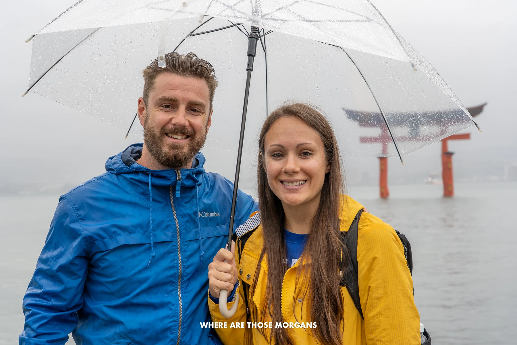 Mark and Kristen from Where Are Those Morgans standing together under a clear umbrealla in bright blue and yellow coats with Itsukushima Shrine on the water behind during a rainy day in Hiroshima Japan
