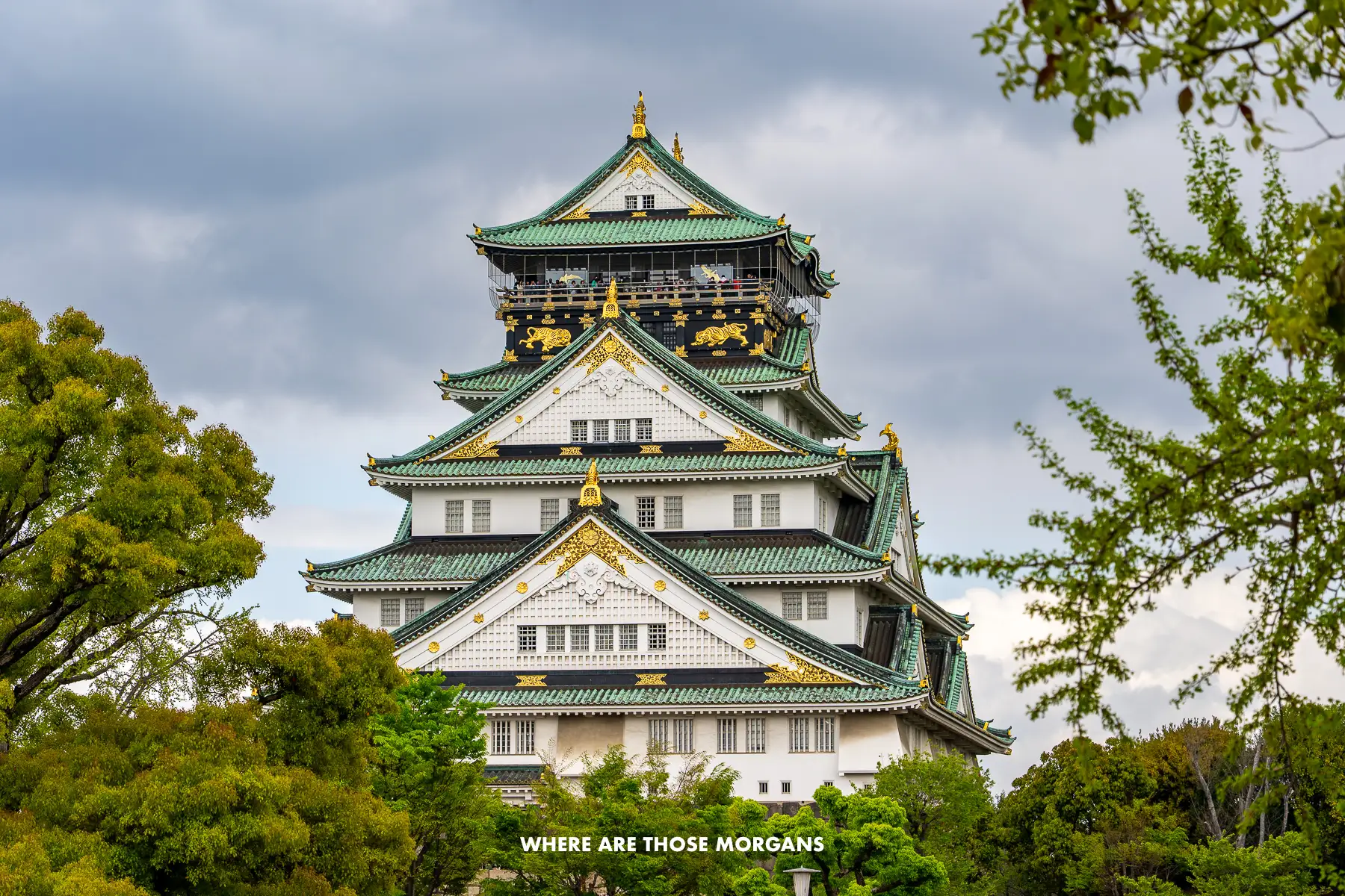 Photo of the green and white Osaka Castle from a middle distance behind trees and below a cloudy sky