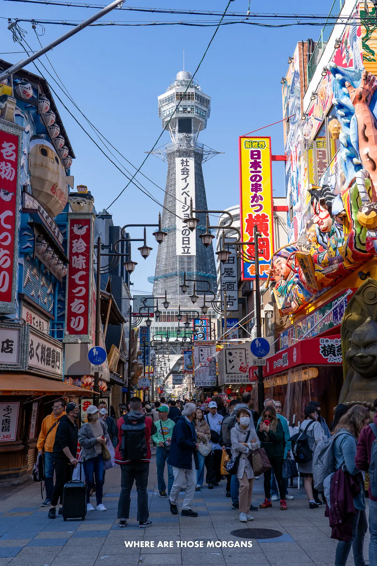 Crowds of people walking through a narrow street flanked by colorful building facades, with a grey tower in the background