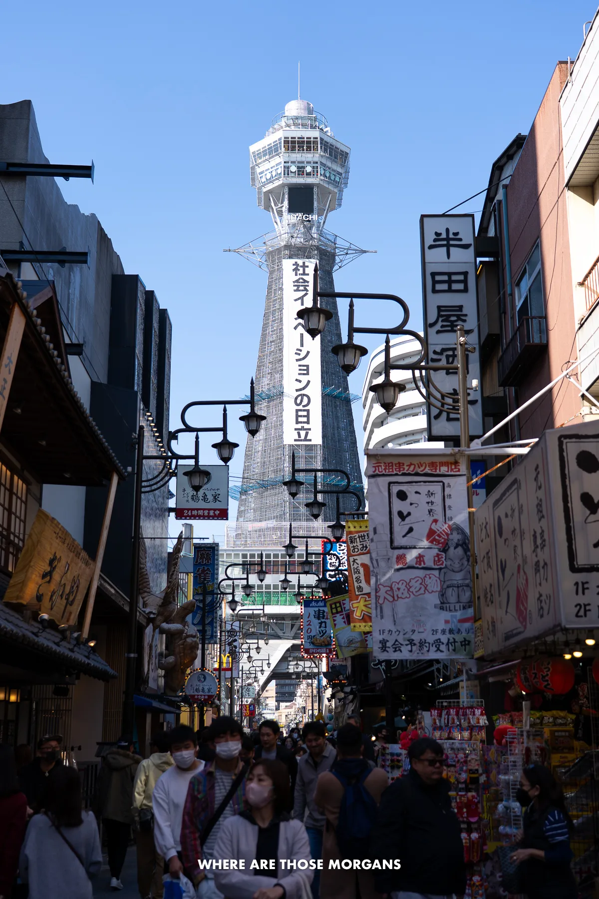 Crowds of people walking through a narrow street lined with eateries and shops, and a tall tower bursting up behind
