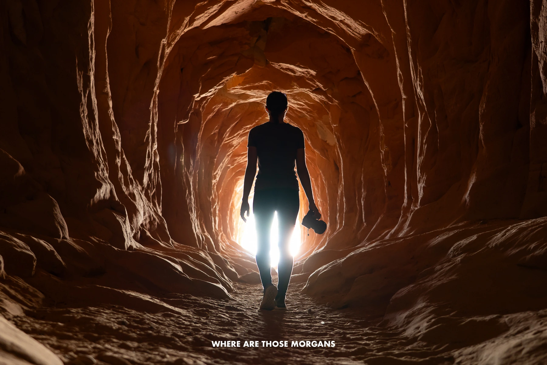 Kristen from Where Are Those Morgans silhouetted holding a camera inside a shadowed sandstone tunnel with light bursting in through the end in Utah