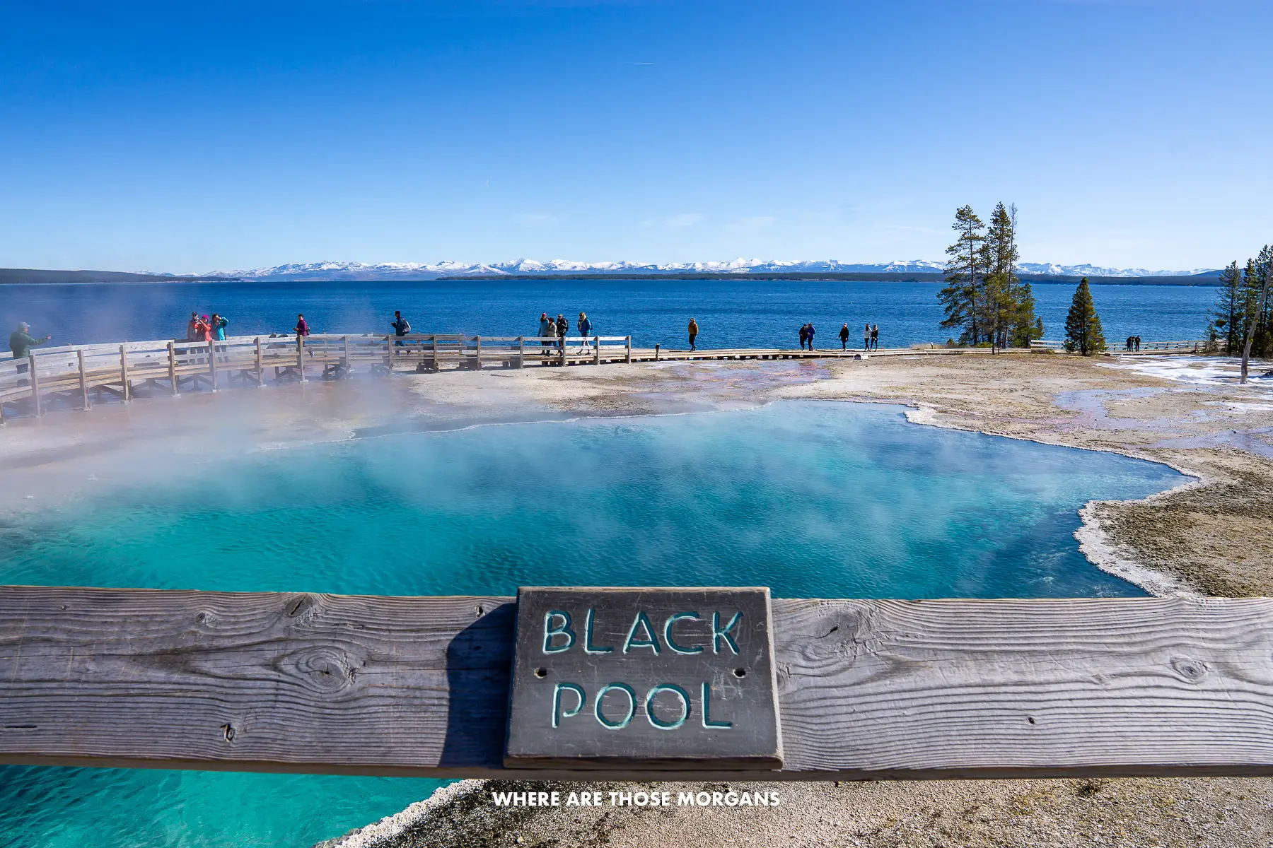 Fence with plaque saying Black Pool with a turquoise colored hot spring leading to a lake and mountains in Wyoming