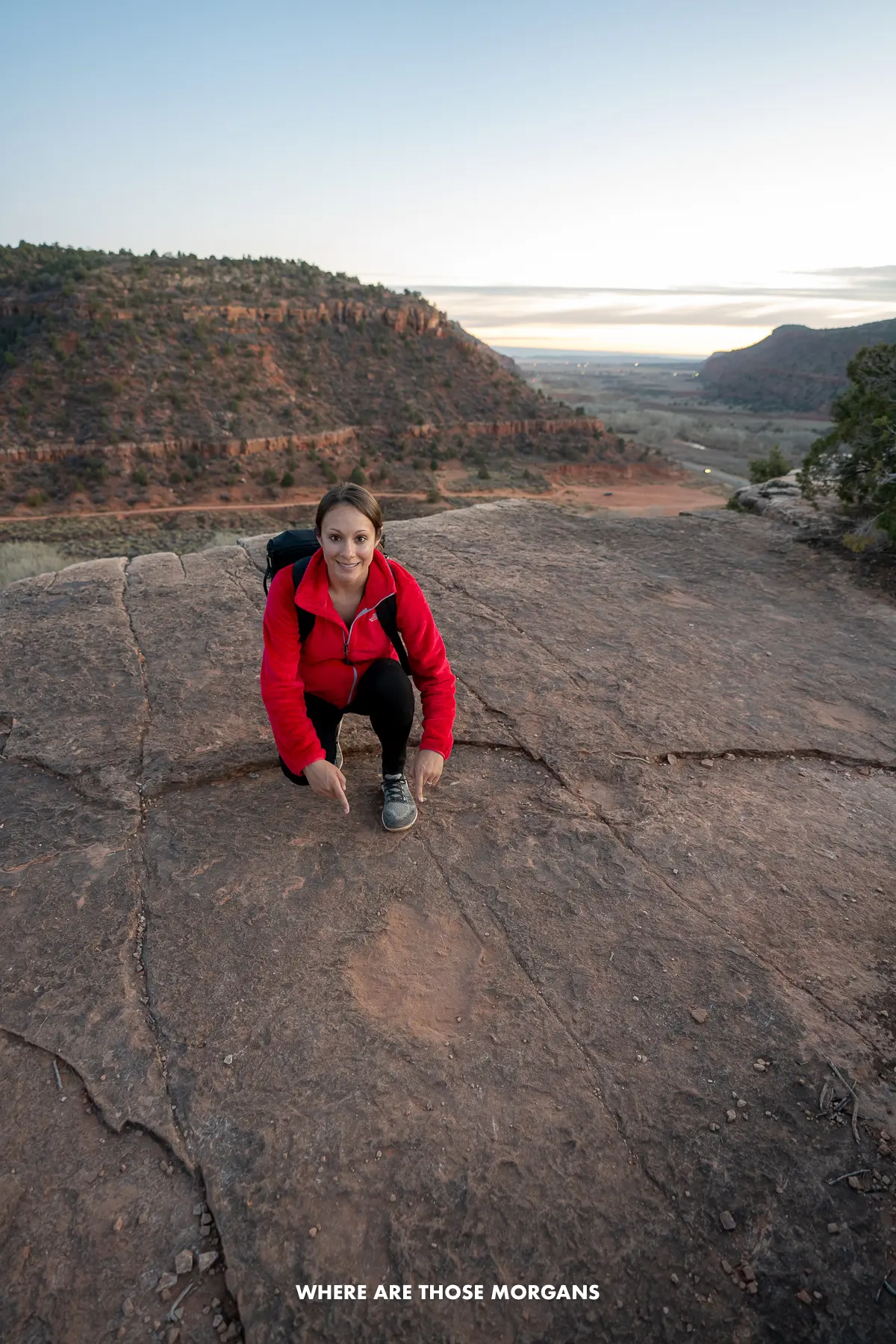 Kristen from Where Are Those Morgans crouching a pointing at a dinosaur print on a flat rocky summit of a short hike called Dinosaur Tracks near Kanab