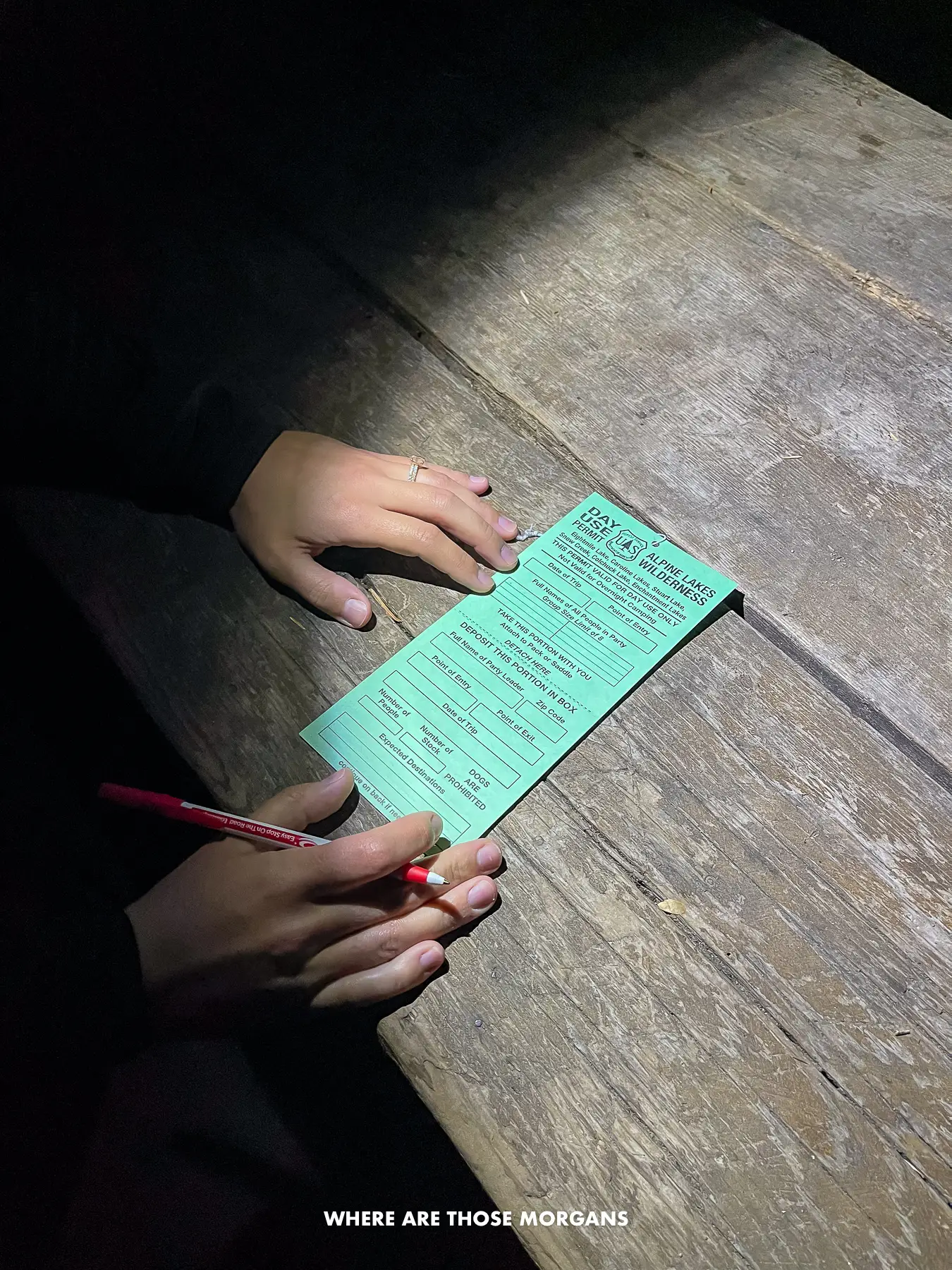 Hands holding a pen and filling in a day use permit on a wooden bench lit by a headlamp light at Stuart/Colchuck Trailhead