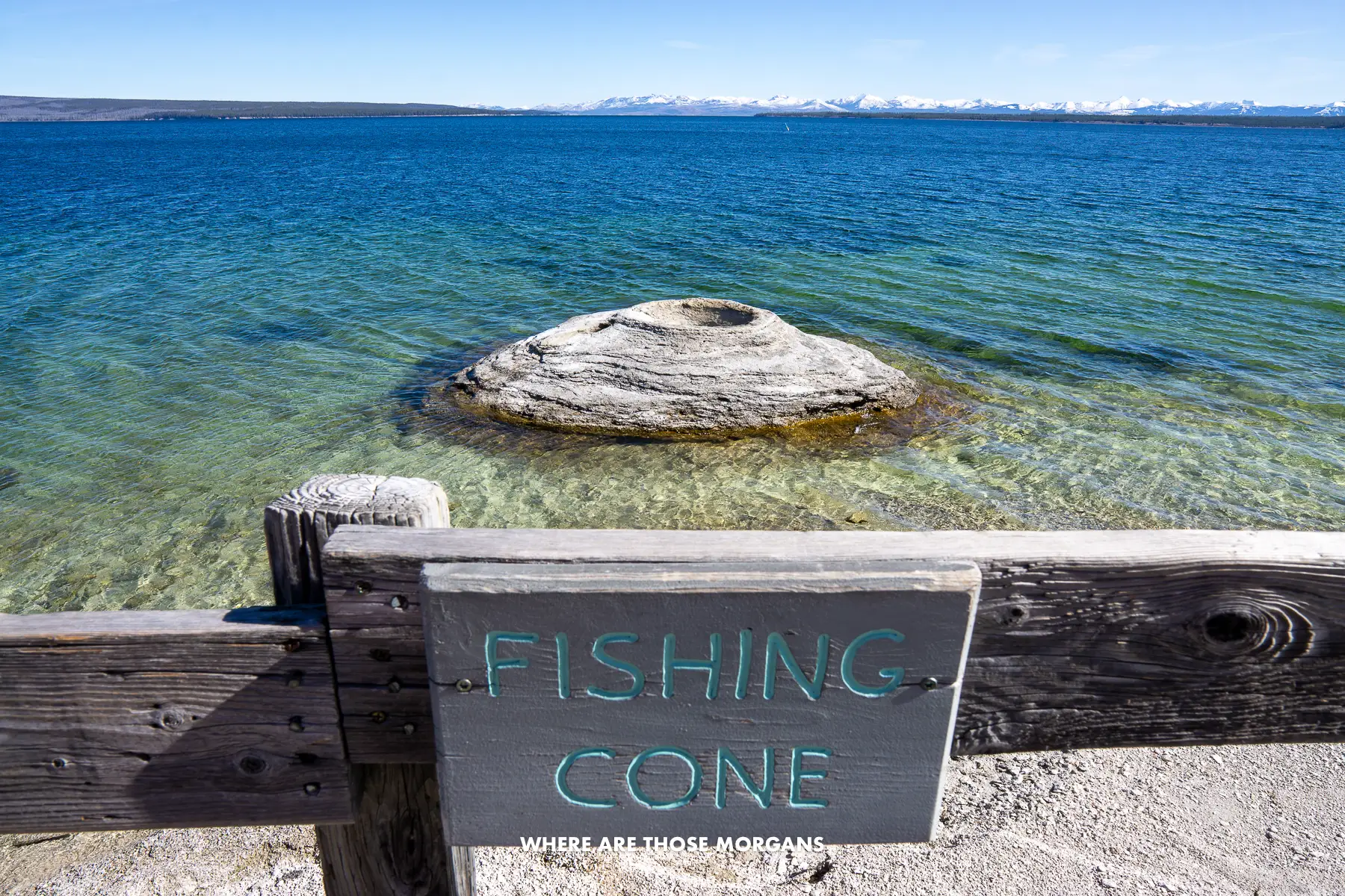 Small Yellowstone geyser in shallow water in a lake with a wooden fence and sign saying Fishing Cone