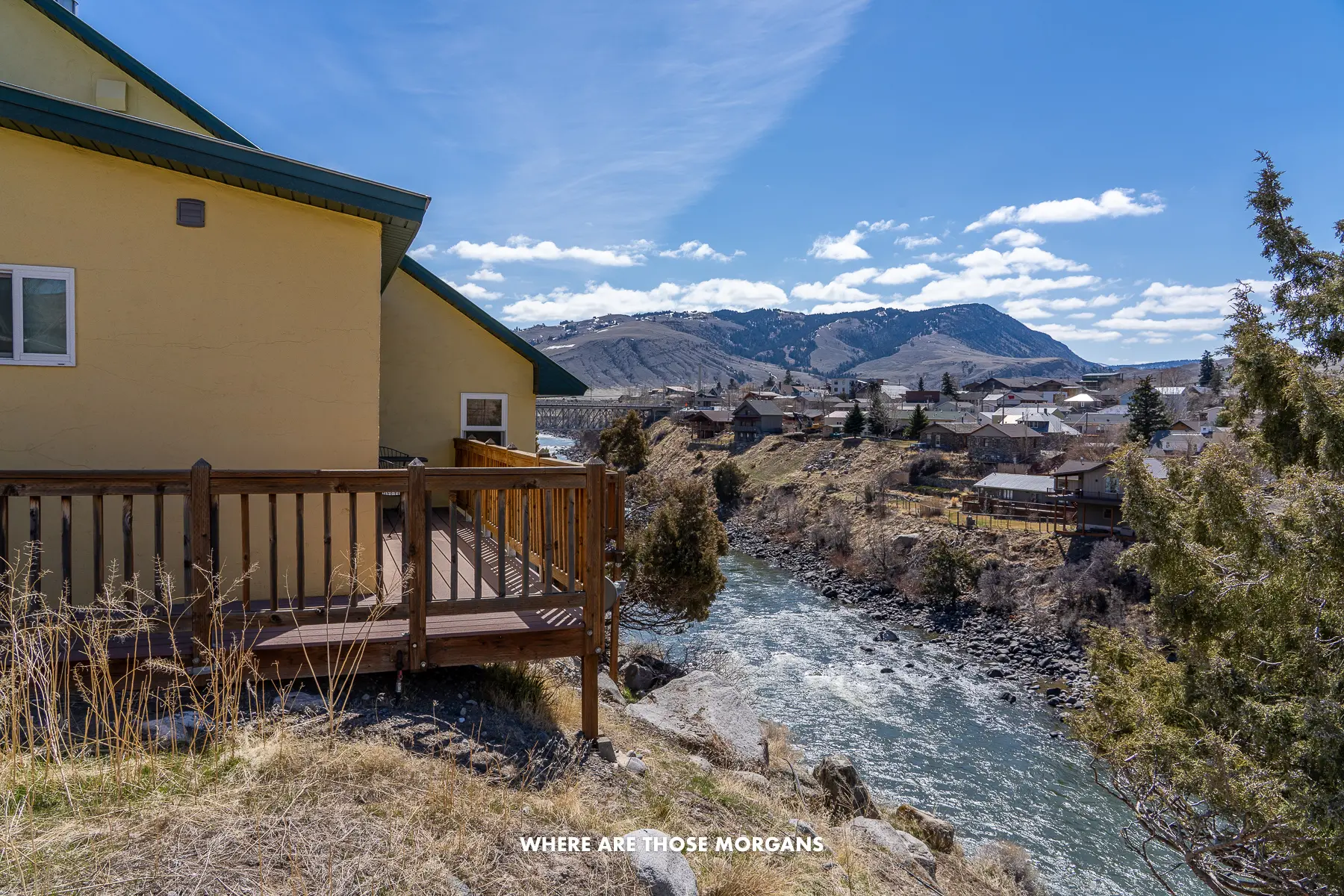 View of a yellow cottage with wooden balcony overlooking a river and rolling hills on a clear day in Montana