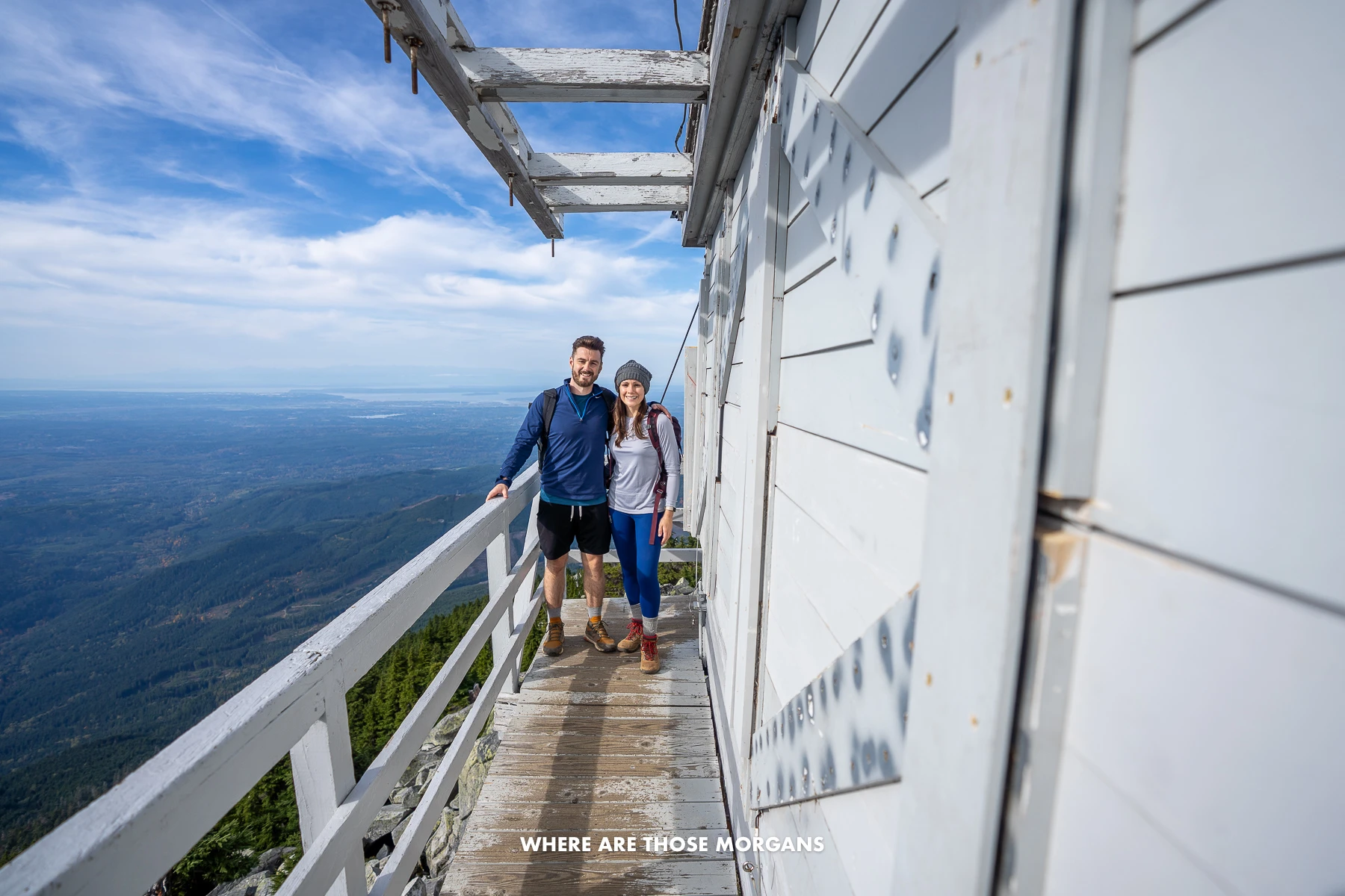 Mark and Kristen from Where Are Those Morgans standing together on a wooden fire tower outer balcony at the summit of Mt Pilchuck in Washington with far reaching views to the side
