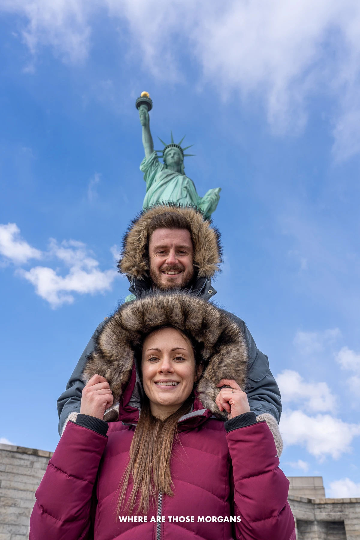 Mark and Kristen from Where Are Those Morgans taking a perspective photo with winter coat hoods up in a line with the Statue of Liberty at the top