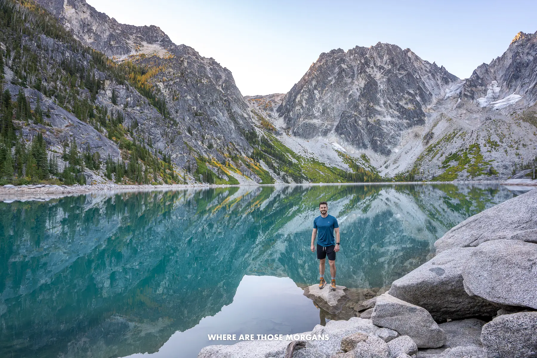 Mark from Where Are Those Morgans stood in shorts and t-shirt on a rock on the shoreline of Colchuck Lake surrounded by tall rugged mountains on The Enchantments hike in Washington at dawn