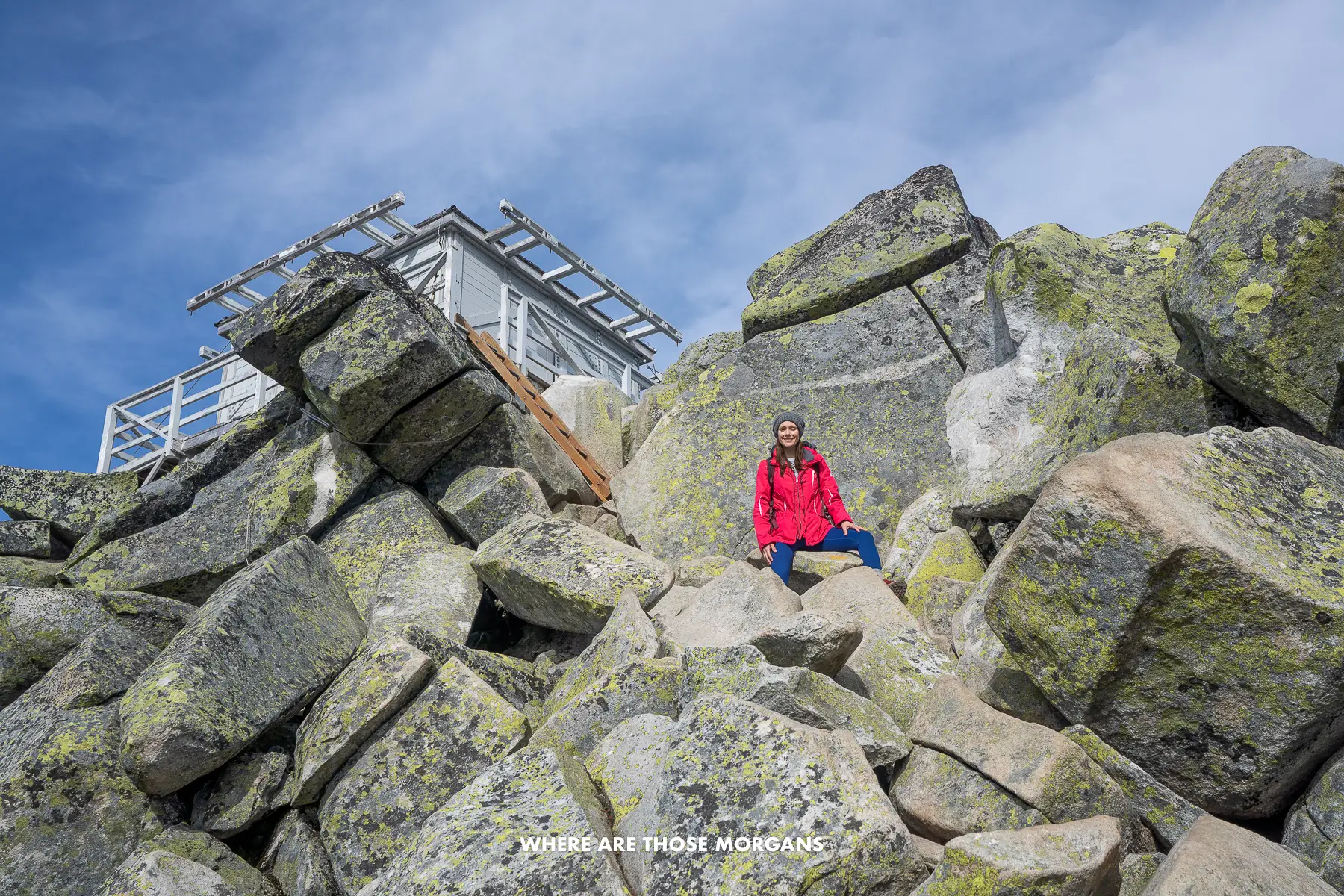 Kristen from Where Are Those Morgans standing on massive boulders with a wooden ladder and fire lookout above at the summit of a trail in Washington