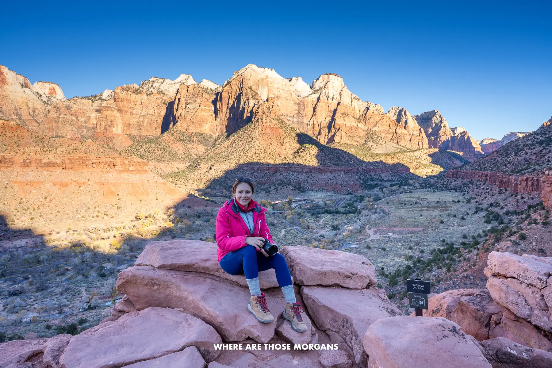Kristen from Where Are Those Morgans sat on red rocks at the summit of a hike in Springdale Utah with sunrise views over red rocks glowing