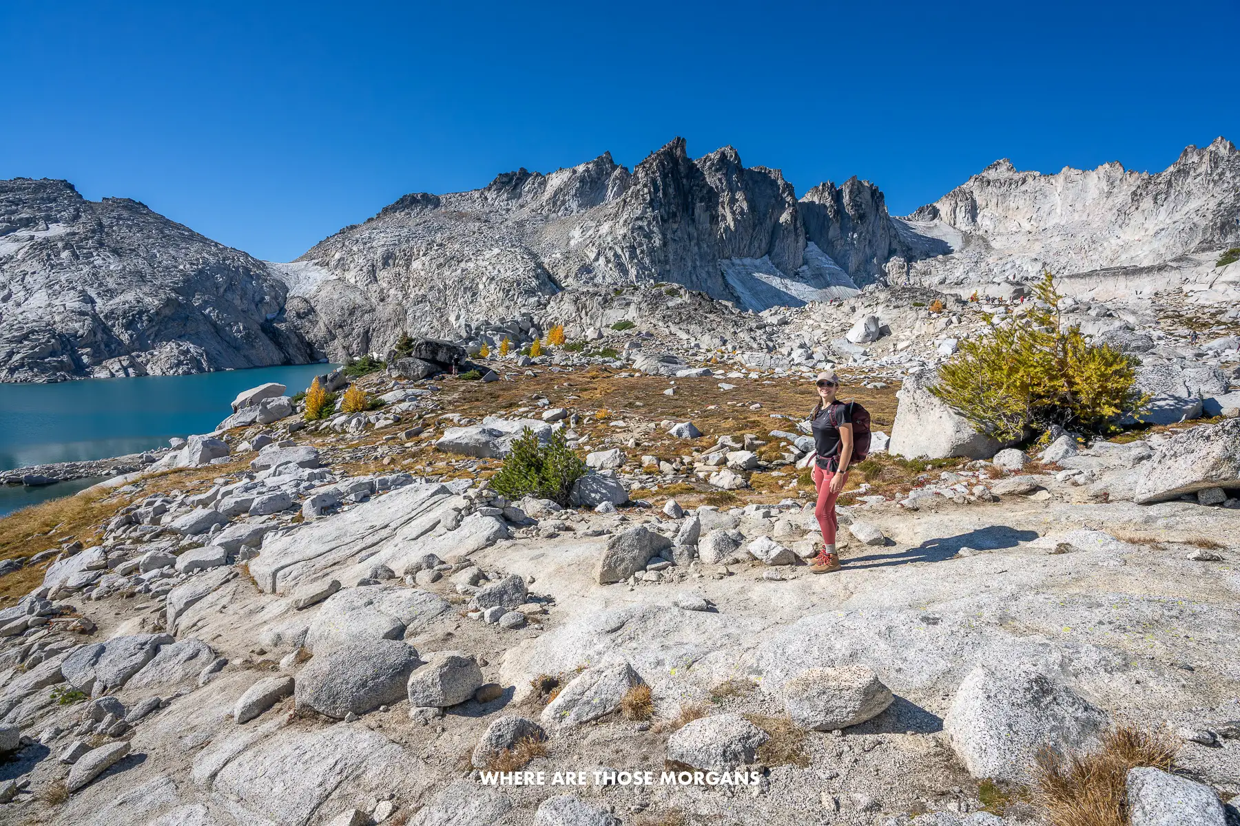 Kristen from Where Are Those Morgans walking on a rocky trail with a lake and rugged granite mountains behind under a deep blue sky