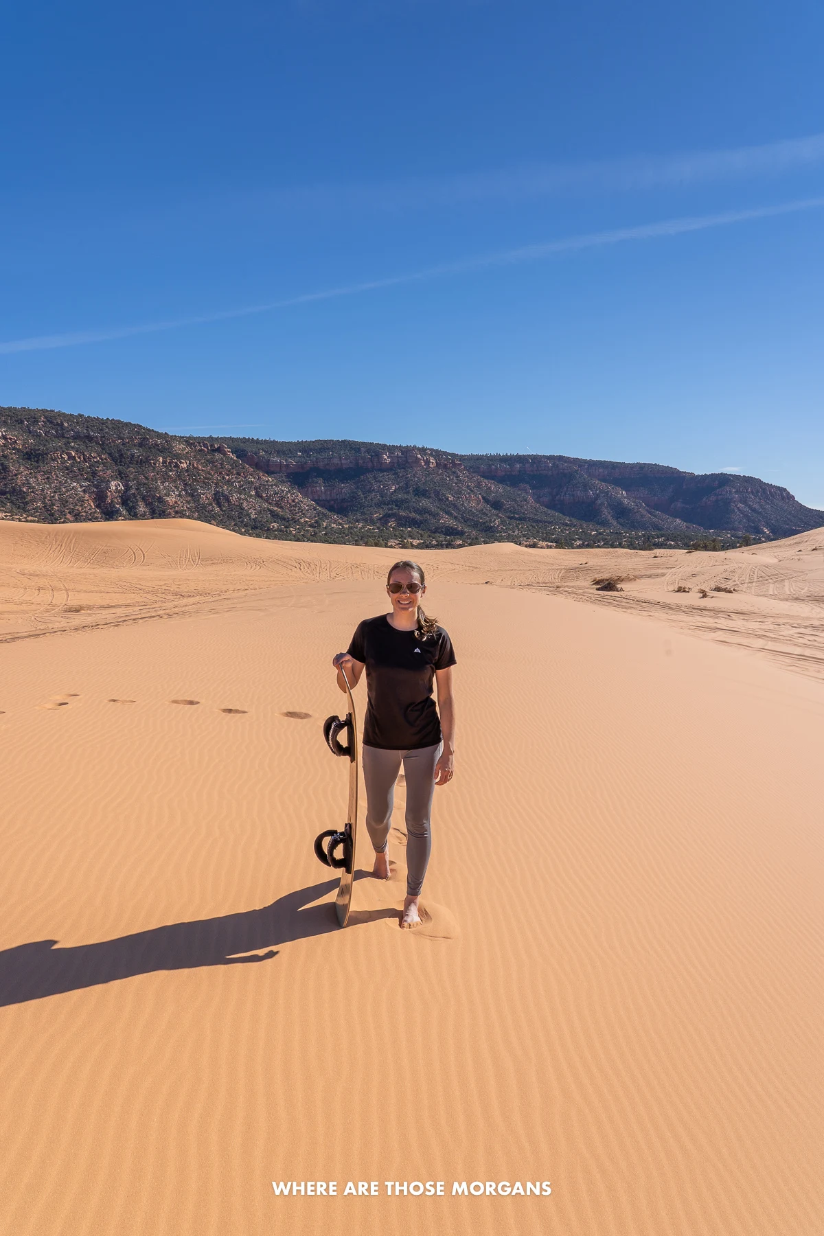 Kristen from Where Are Those Morgans walking on a massive sand dune holding a sand board under a clear blue sky 