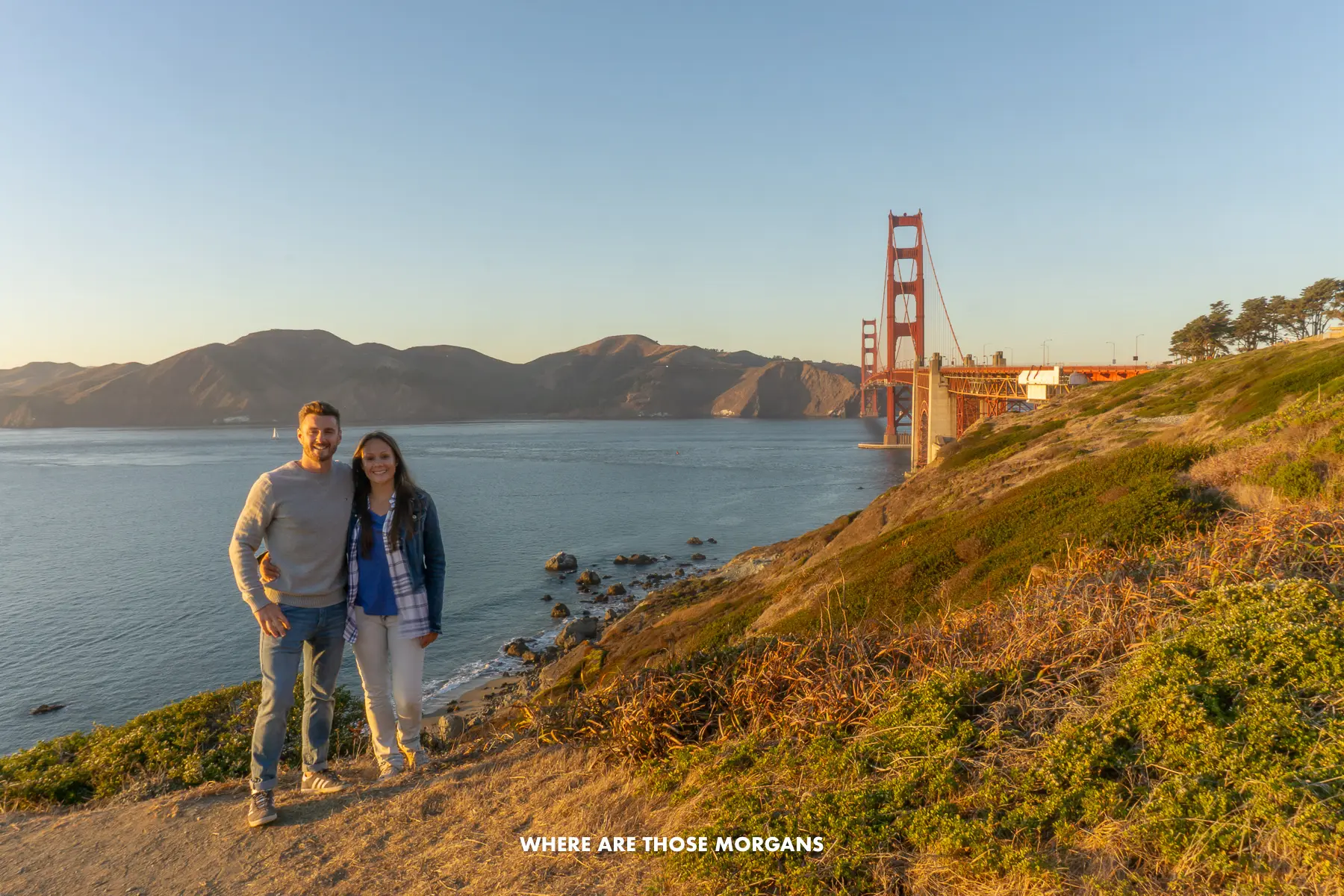 Mark and Kristen from Where Are Those Morgans standing together for a photo on the side of a cliff at sunset with views over the Golden Gate Bridge and San Francisco Bay