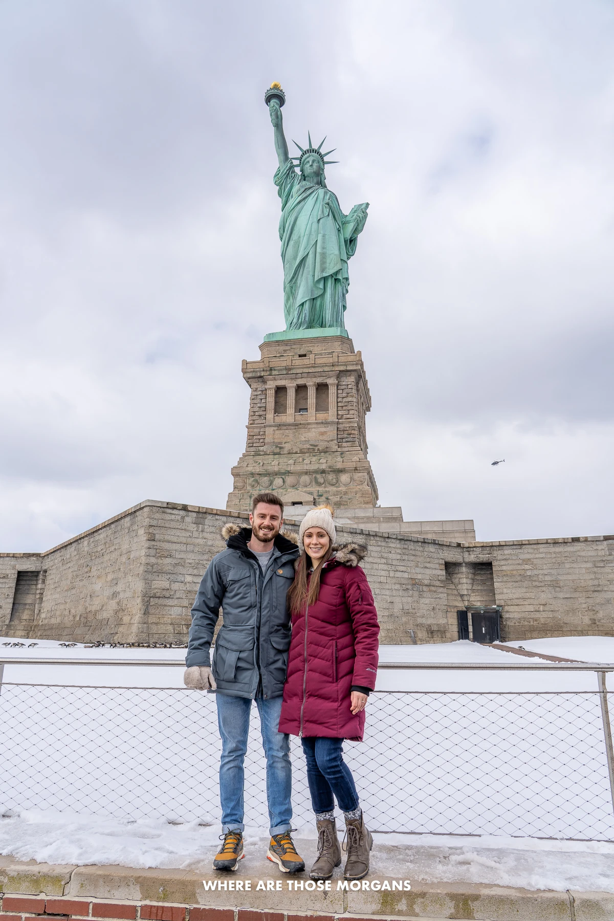 Mark and Kristen from Where Are Those Morgans standing together on Liberty Island with snow on the ground and the Statue of Liberty right behind