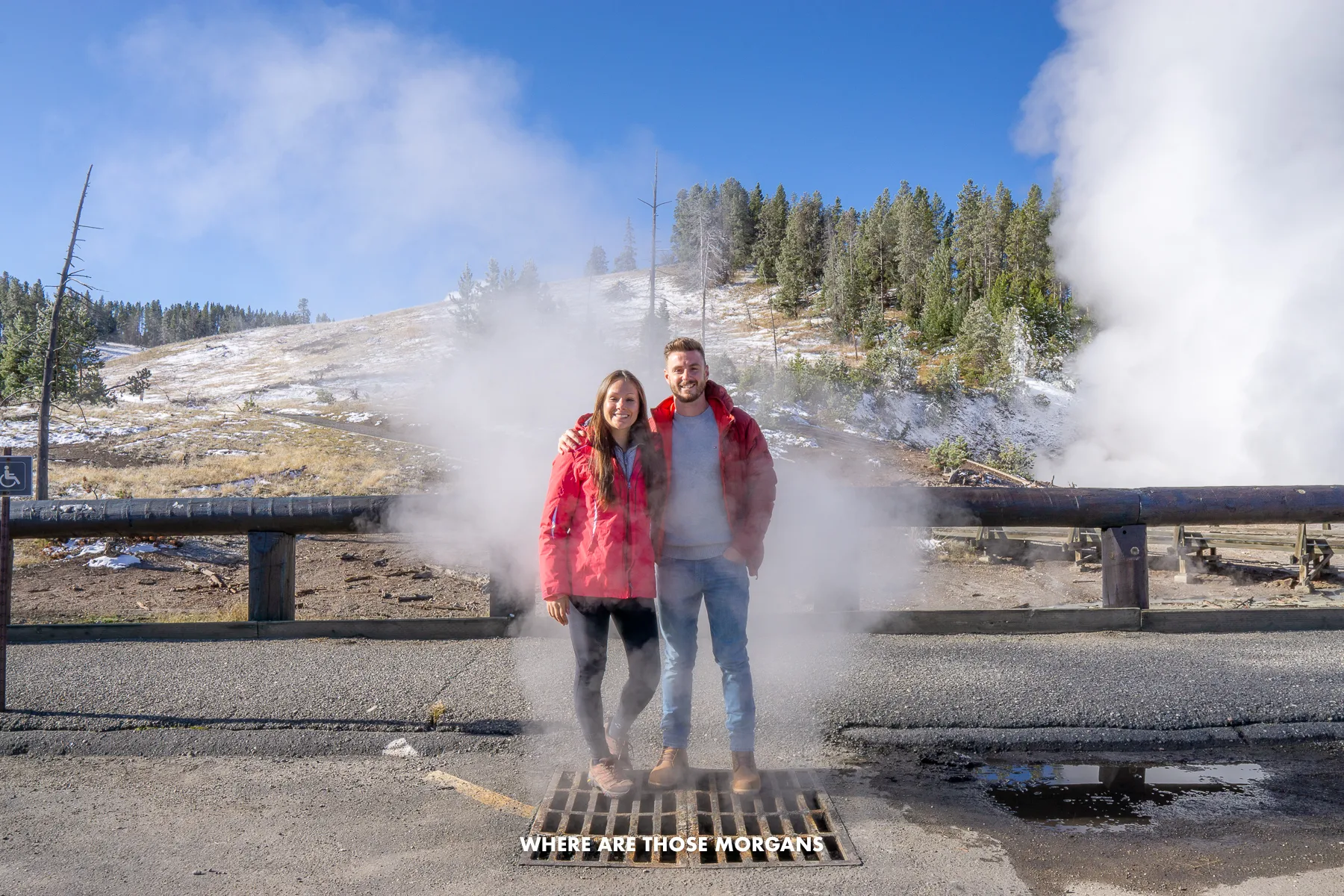 Mark and Kristen from Where Are Those Morgans standing together on a grate in a parking lot with billowing geysers and mud pots behind in Yellowstone National Park