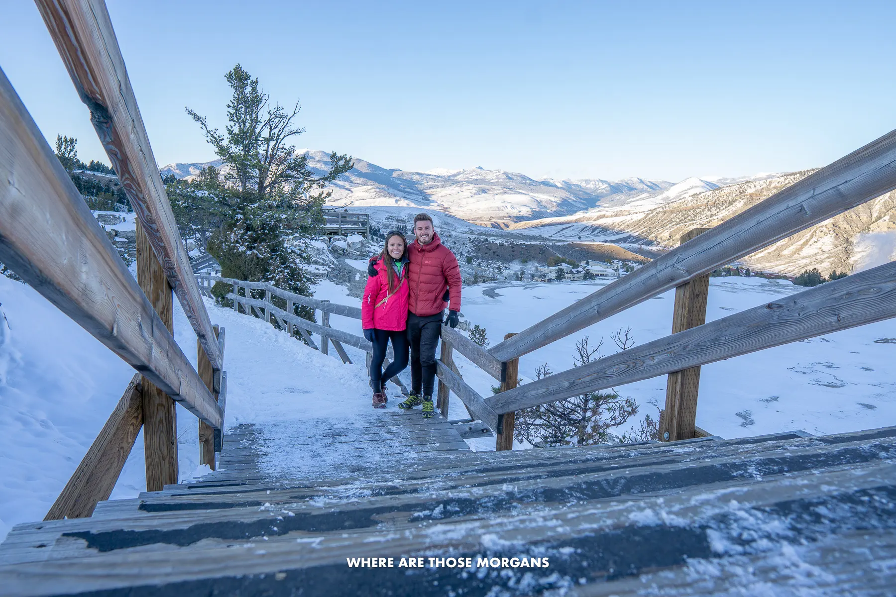 Mark and Kristen from Where Are Those Morgans standing together on a wooden boardwalk covered in snow in Yellowstone National Park at sunrise with distant mountain views