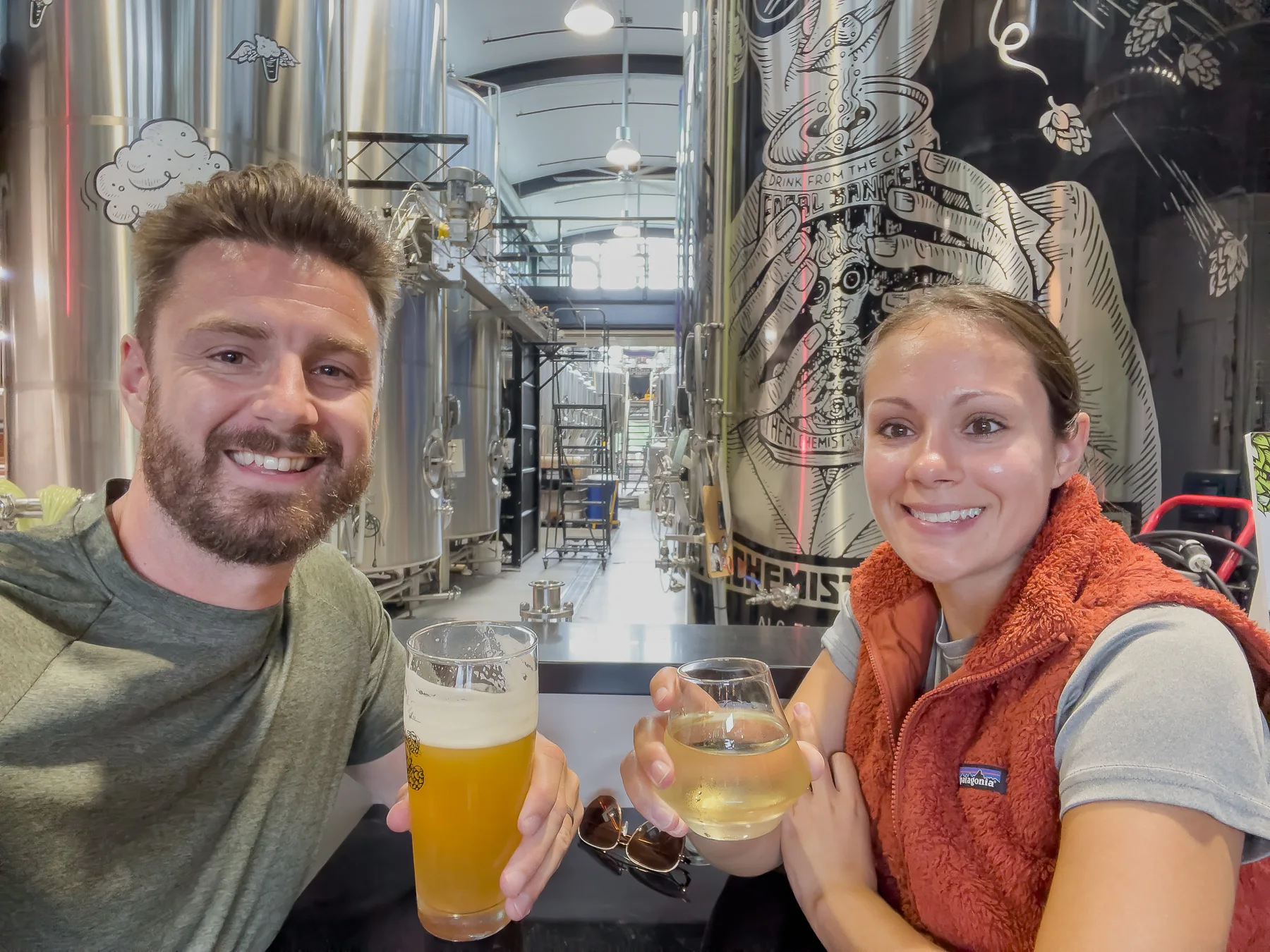 Photo of Mark and Kristen Morgan from Where Are Those Morgans with a beer each inside a brewery called The Alchemist in Stowe with huge brewing tanks behind