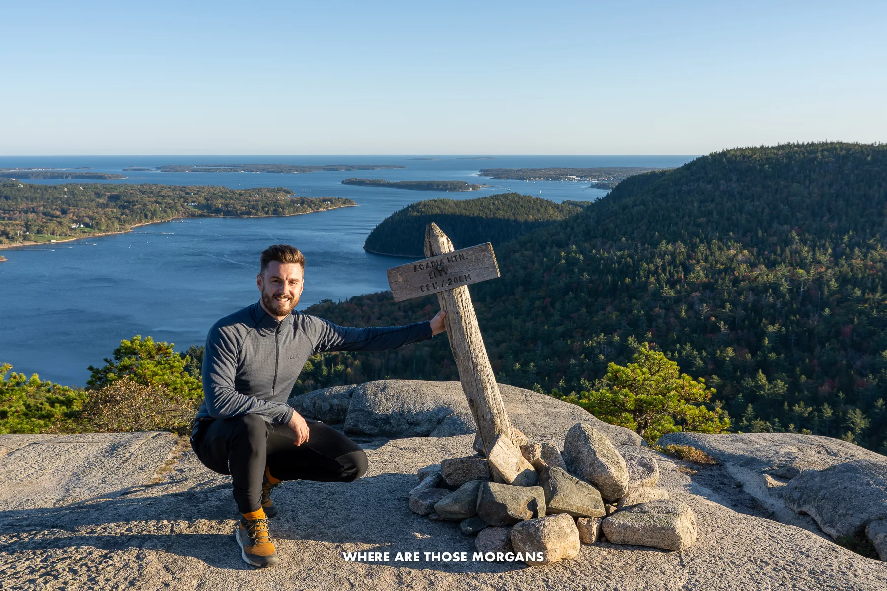 Mark from Where Are Those Morgans crouching with a sign saying Acadia Mountain at the rocky summit with views over lakes behind