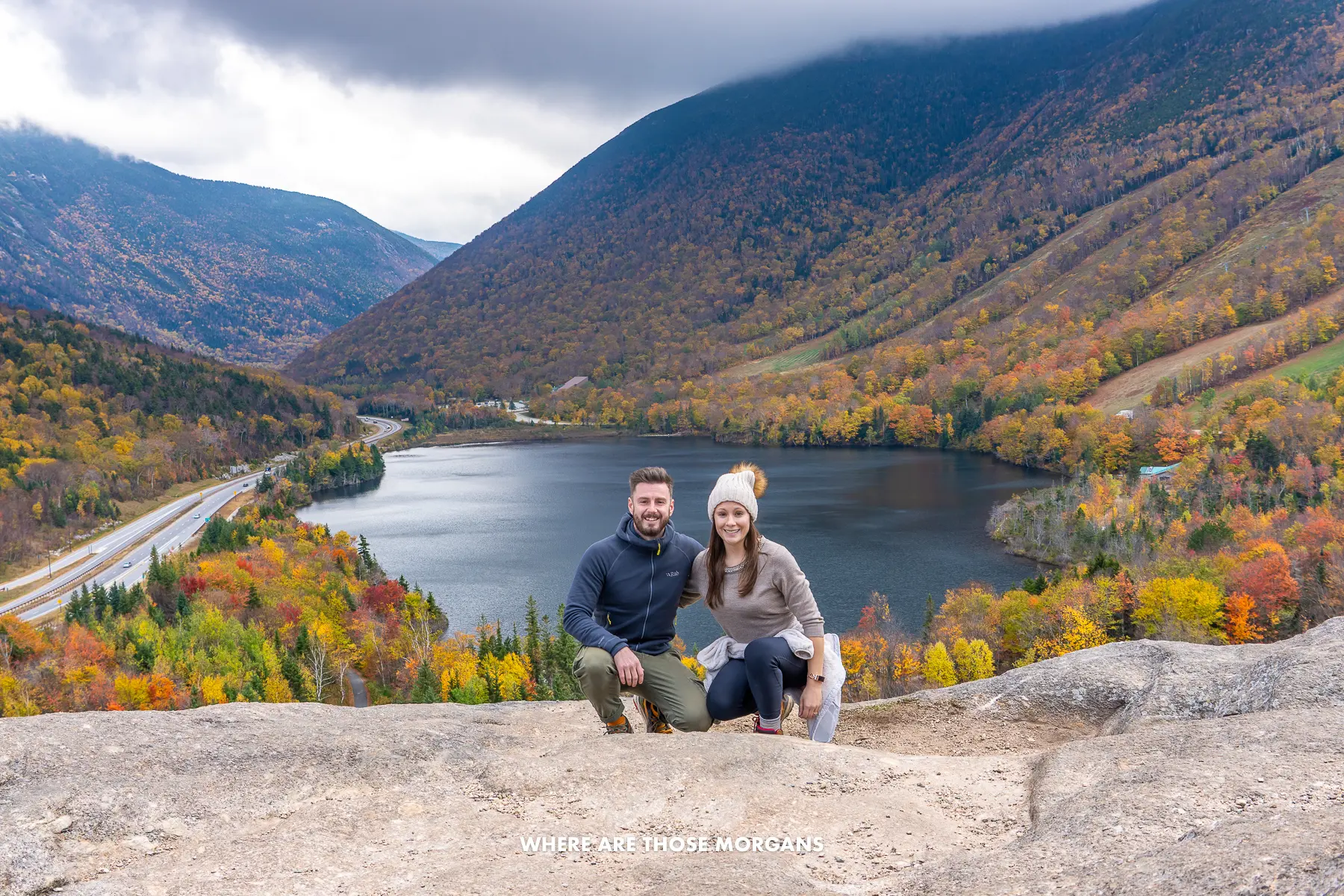 Mark and Kristen from Where Are Those Morgans crouching together for a photo on a rocky summit overlooking a lake and rolling hills covered in trees with colorful leaves in New England in the fall