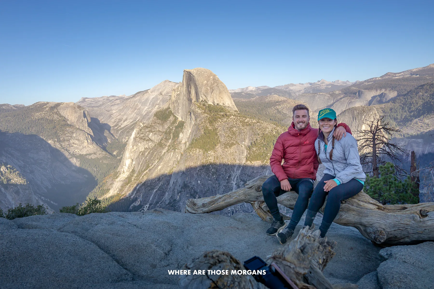 Mark and Kristen from Where Are Those Morgans sitting together on a log with wide open views behind over Half Dome and Yosemite at sunset