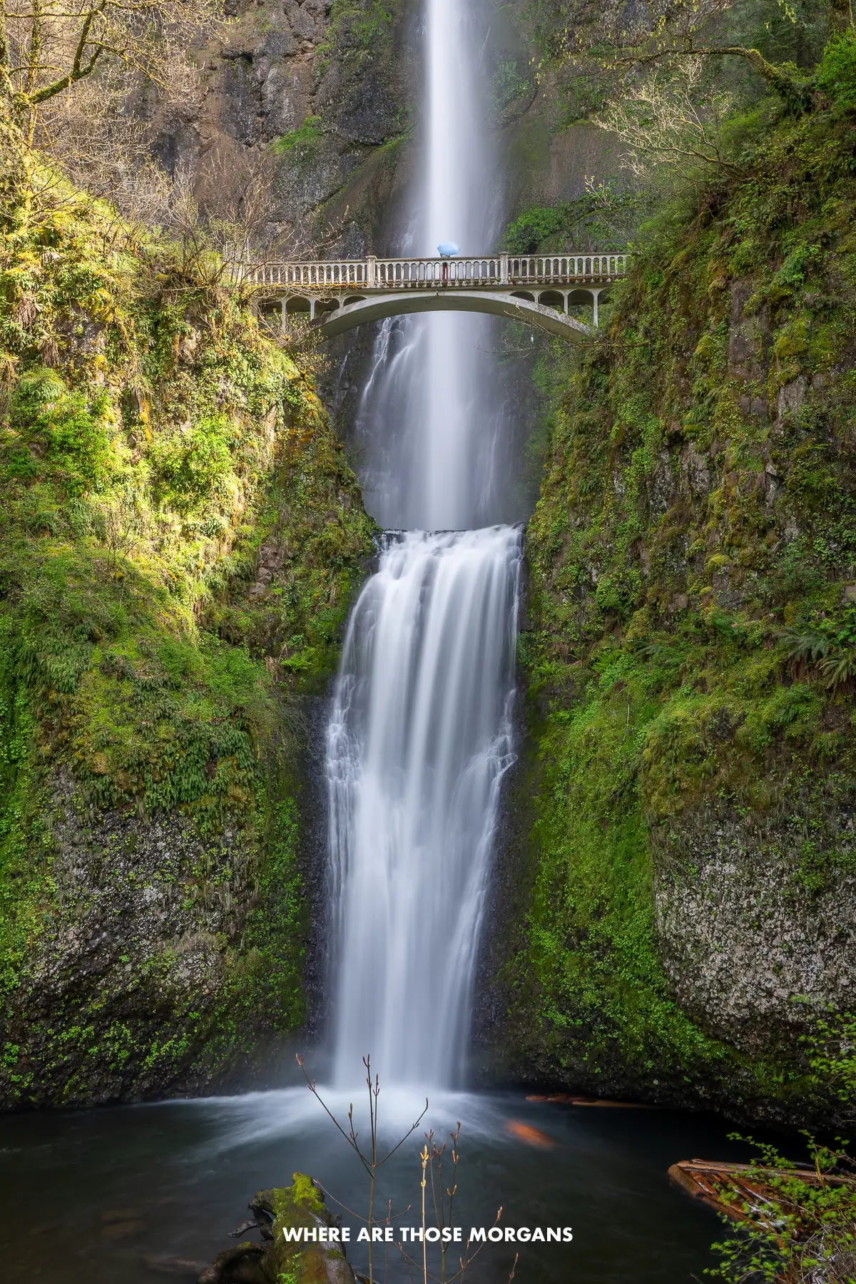 Kristen from Where Are Those Morgans spinning a blue umbrella on a small wooden bridge crossing Multnomah Falls in Oregon