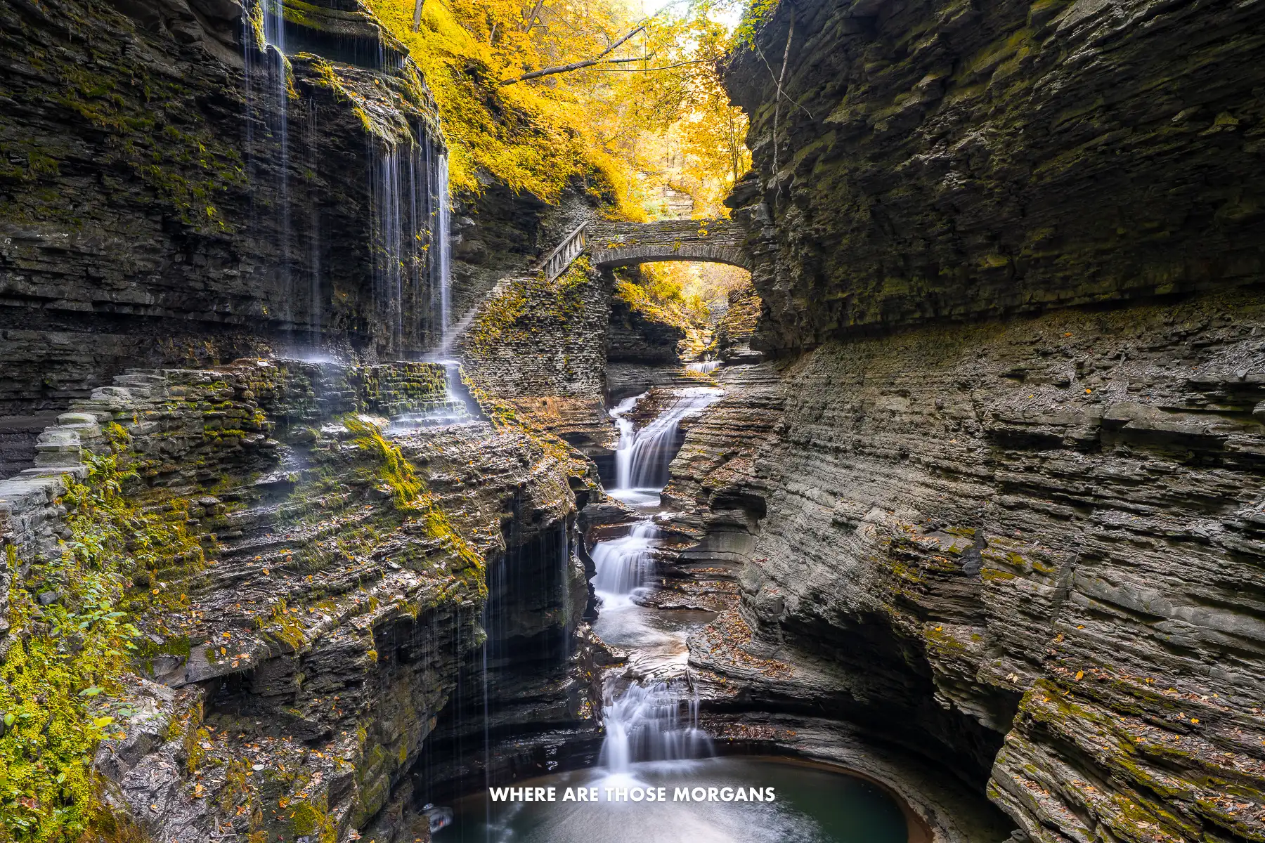 Zig zagging waterfall lightly flowing through a narrow gorge with water falling from above and yellow leaves above a stone bridge in Watkins Glen gorge