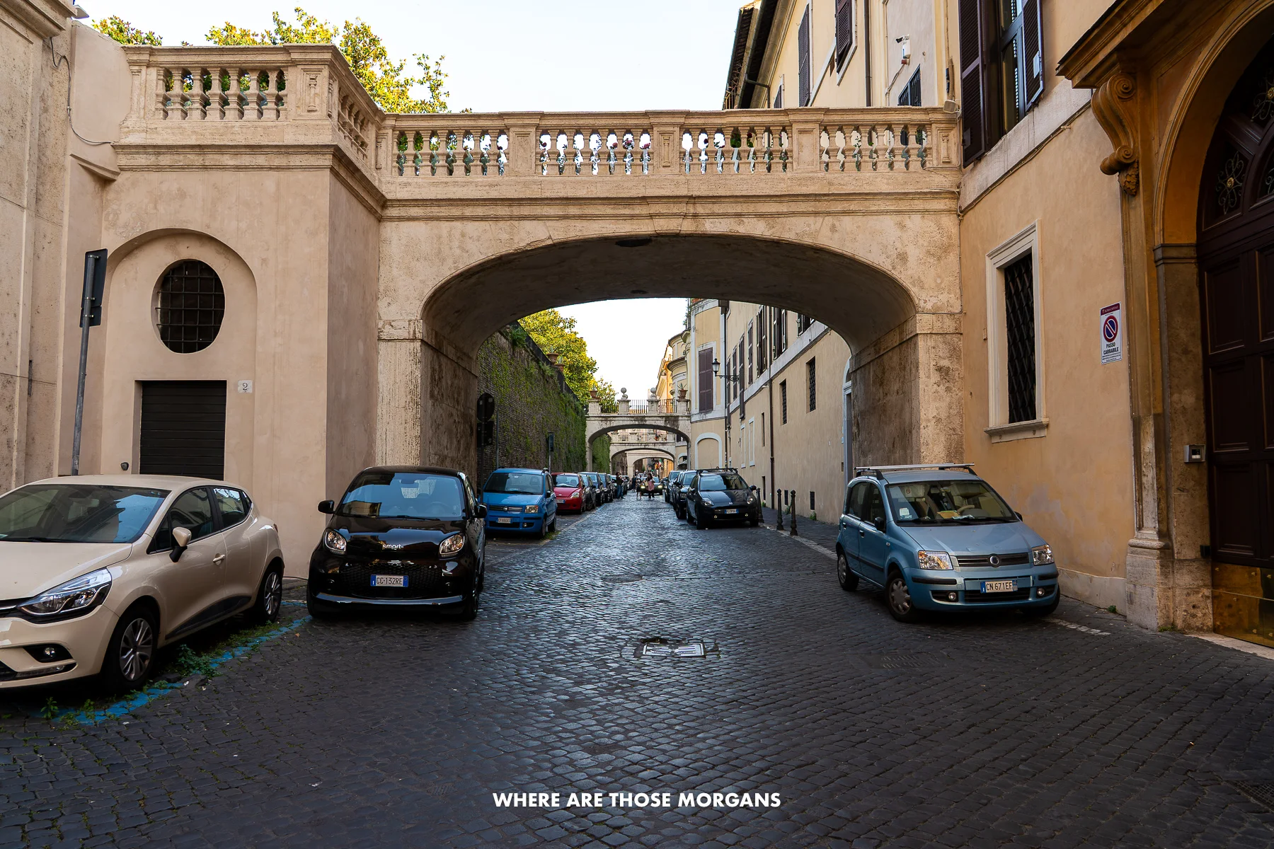 Cobbled road with cars parked and an old small bridge crossing between buildings