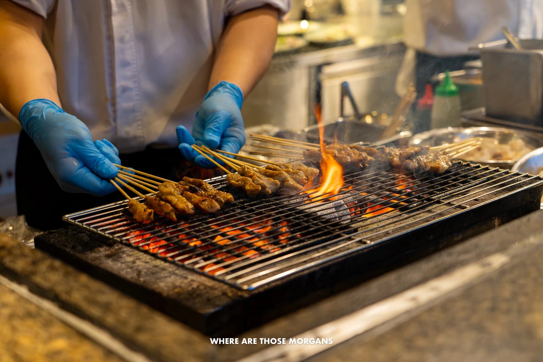 Person with gloves holding chicken satay skewers over a hot grill with flames
