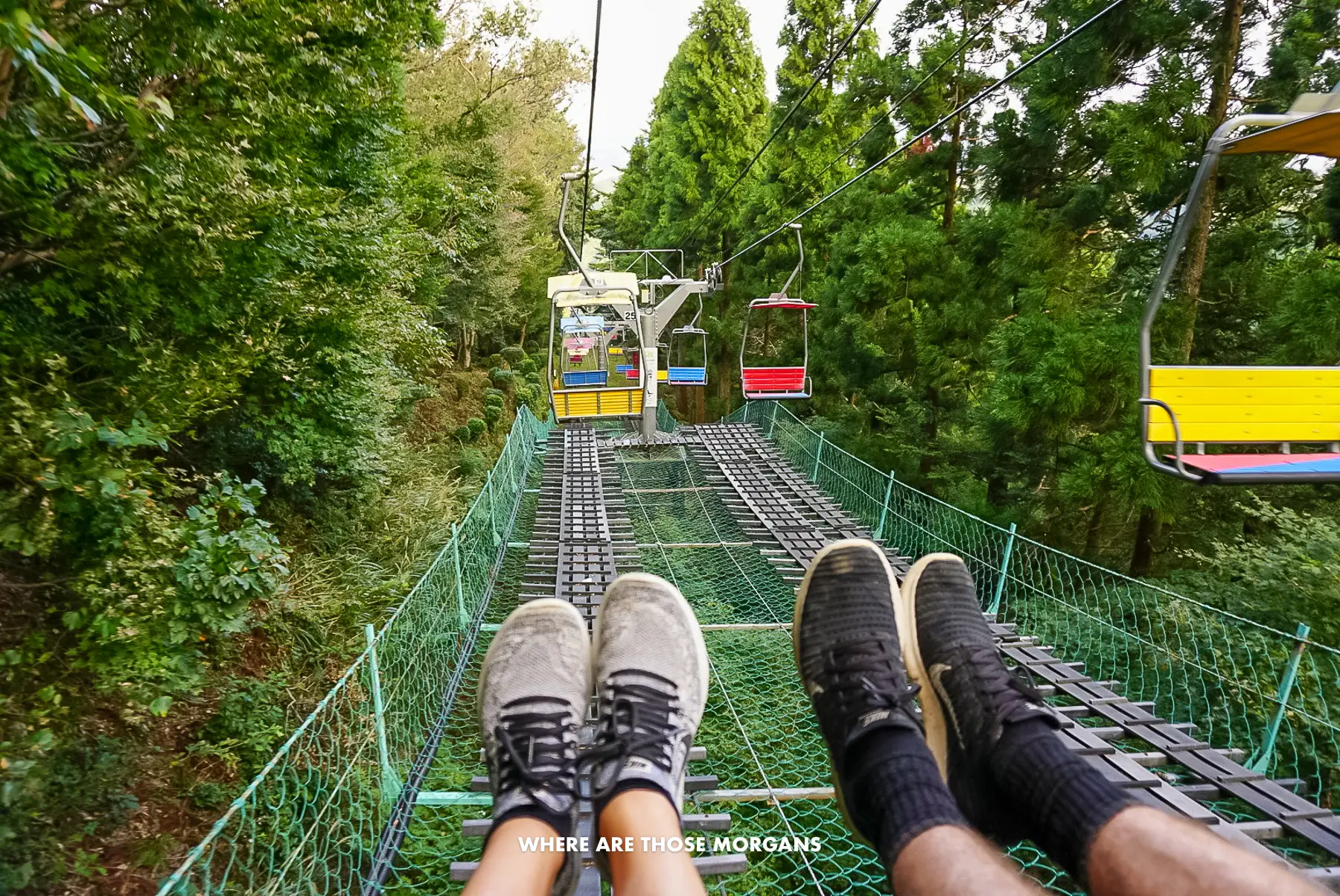 Two pairs of legs and feet with sneakers on a chair lift above a net going through trees