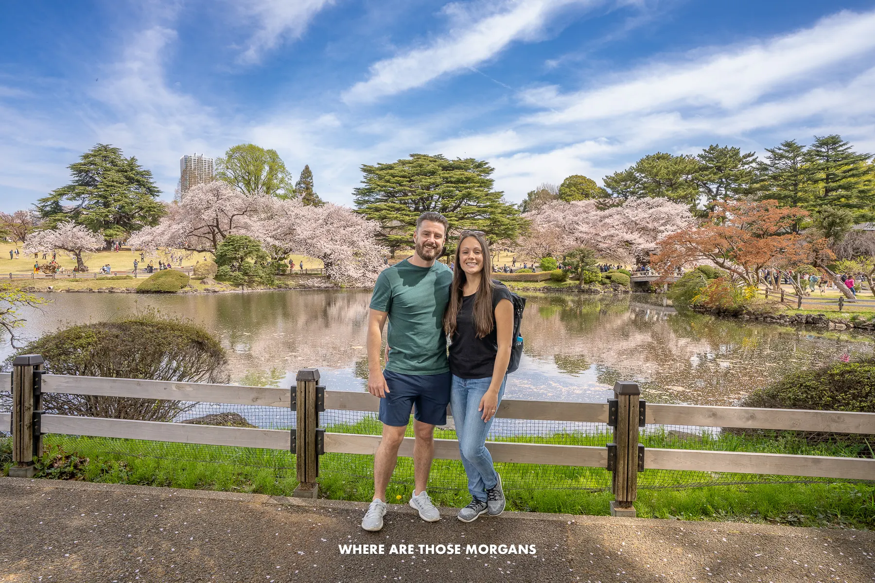 Mark and Kristen from Where Are Those Morgans having a photo taken in Shinjuku National Garden with a small fence, pond and trees behind on a sunny day