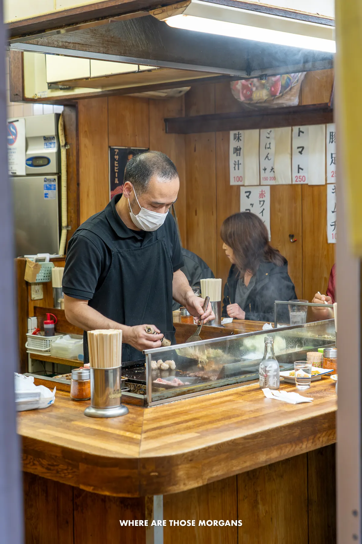 Japanese chef cooking food on a hot plate surrounded by a U-shaped wooden table with people eating