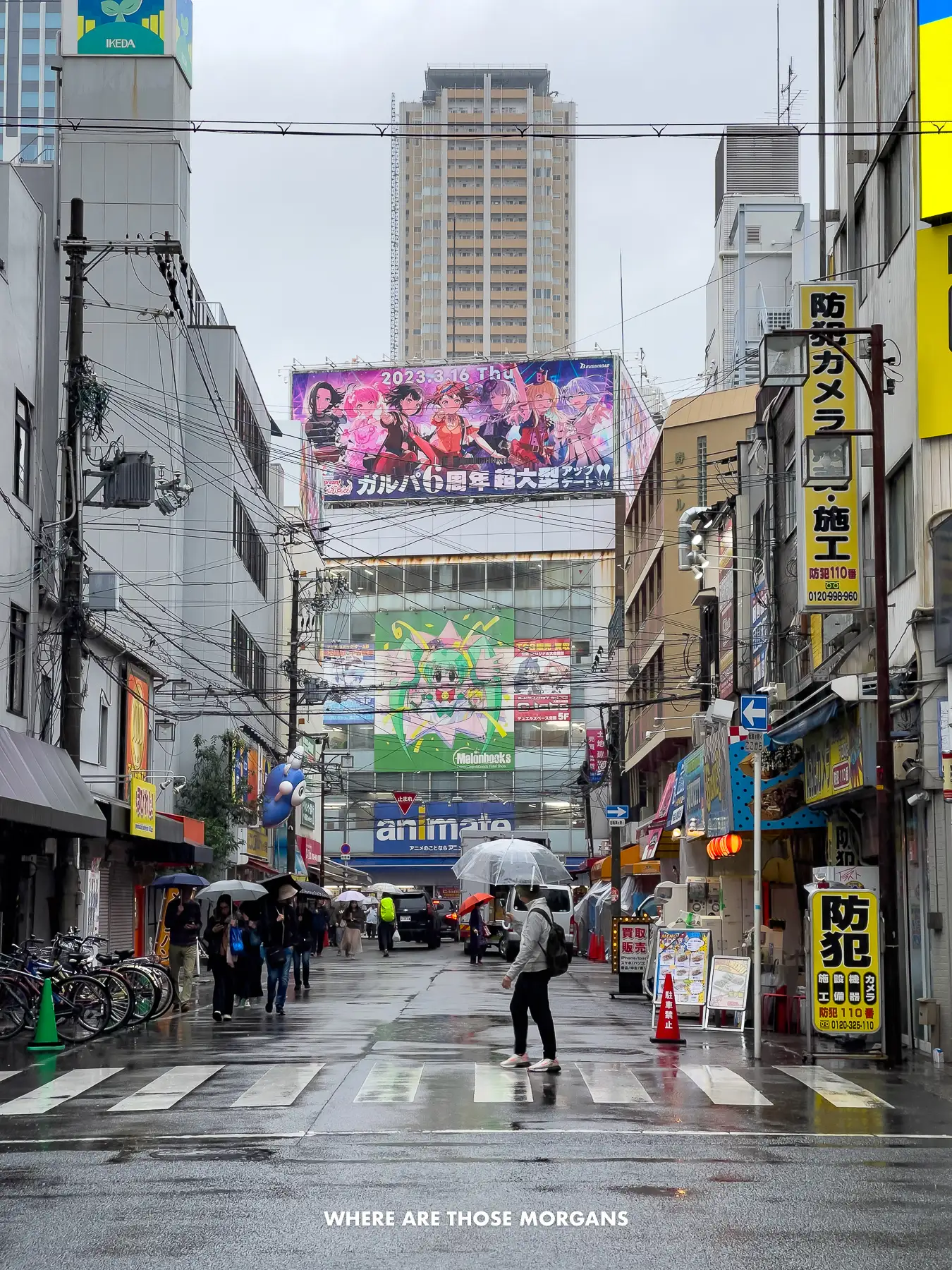 People walking across a road in the rain between tall grey buildings in Osaka
