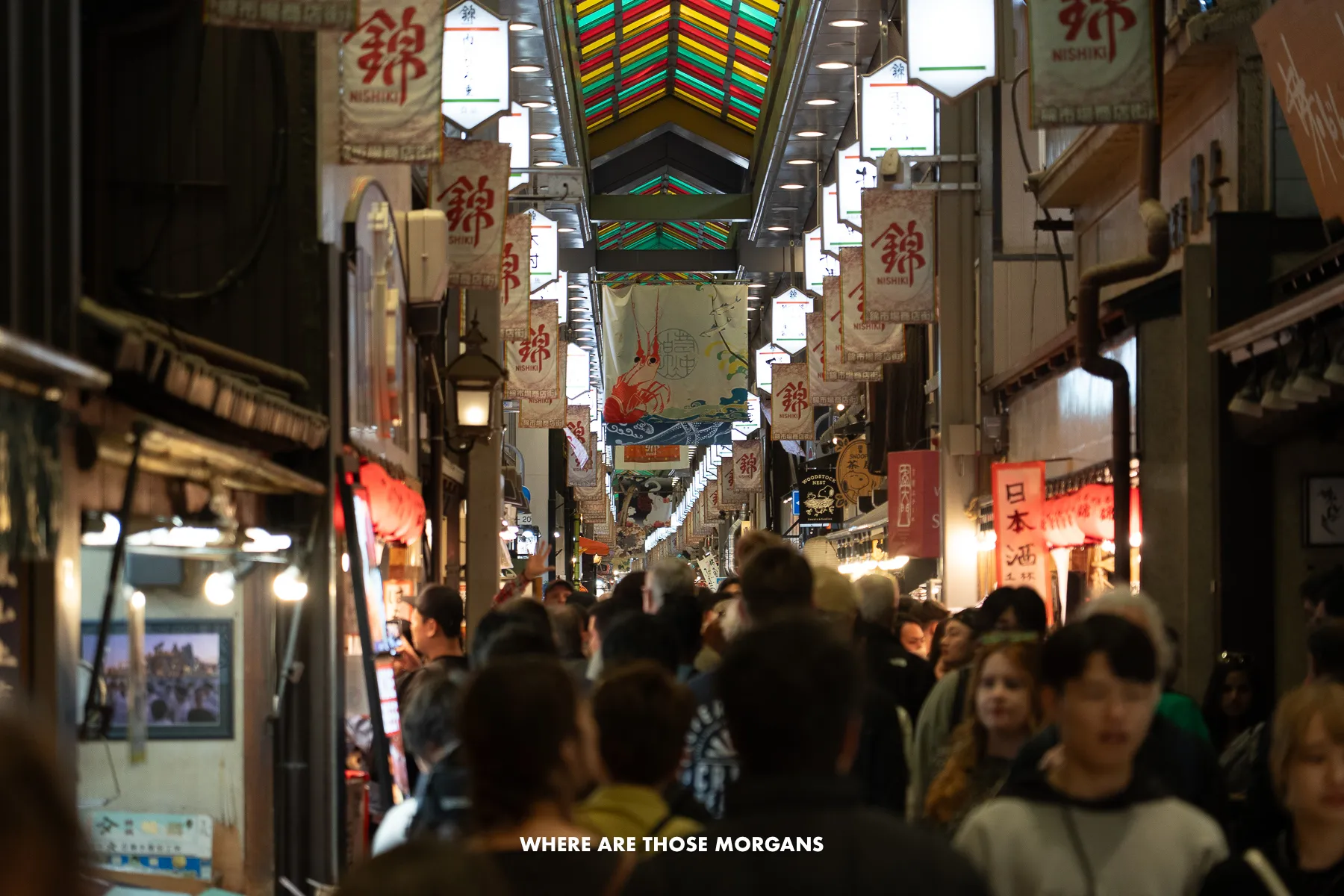 Crowd of people walking down a narrow walkway inside Nishiki Market in Kyoto at night with lights above the stalls