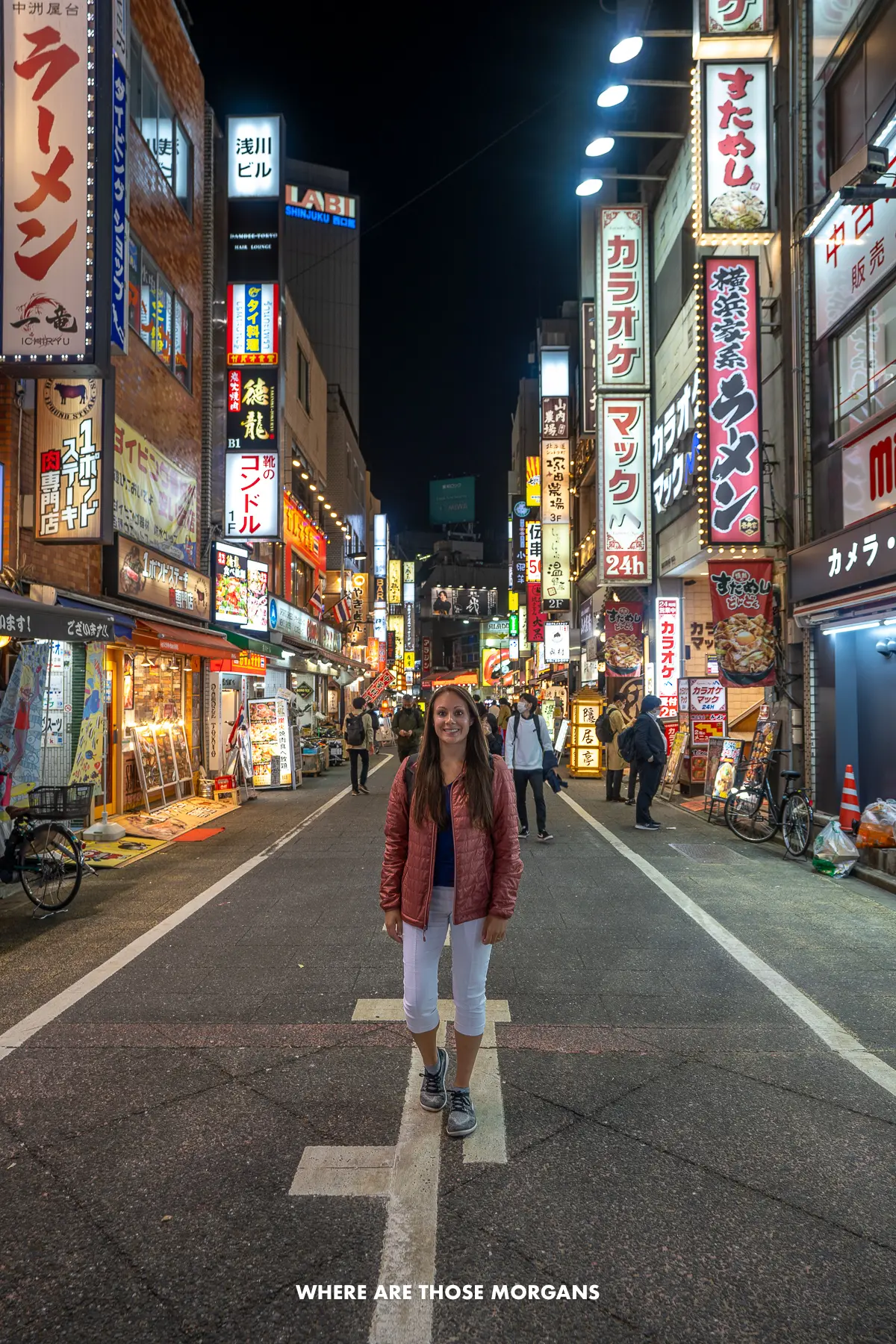 Kristen from Where Are Those Morgans standing in the middle of a narrow street in Shinjuku, Japan at night with neon lit buildings and other people behind