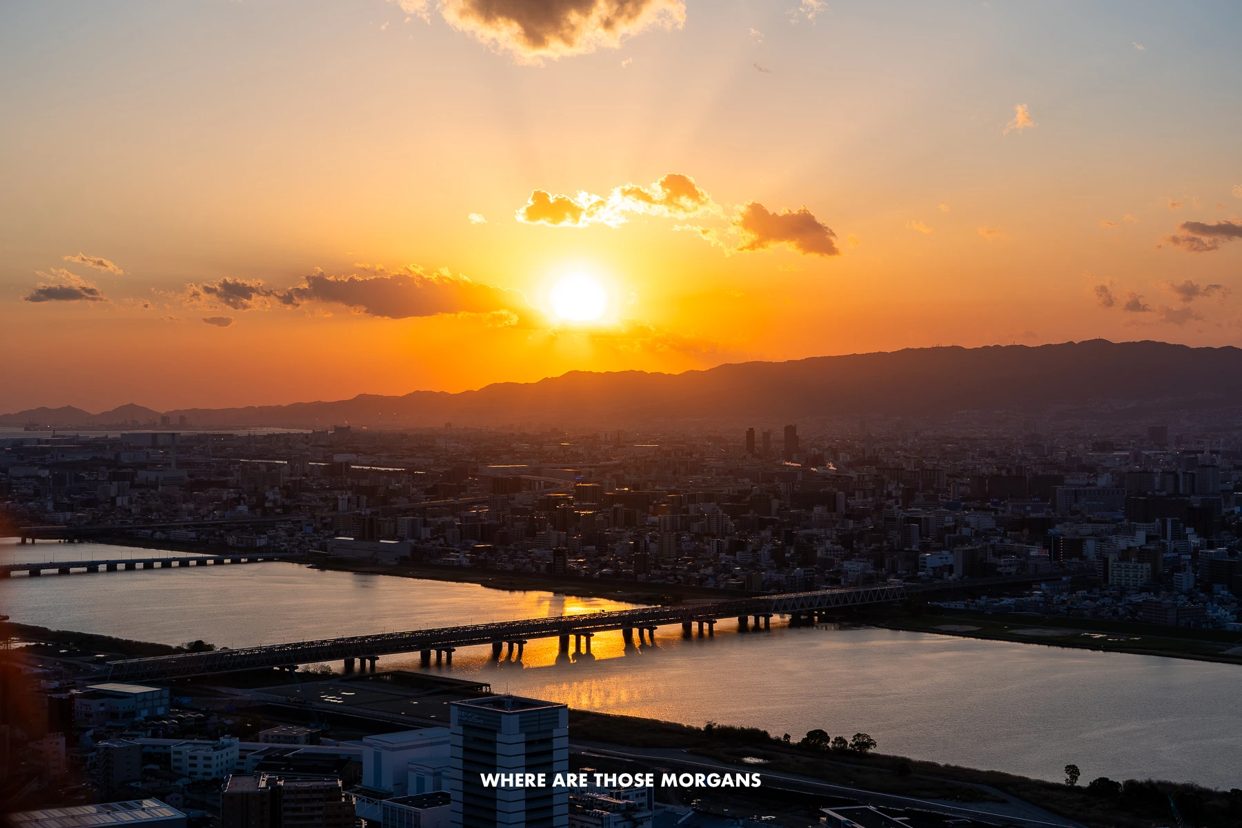 Serene and colorful sunset with light clouds above a silhouetted shallow row of mountains with a river and bridge in the foreground